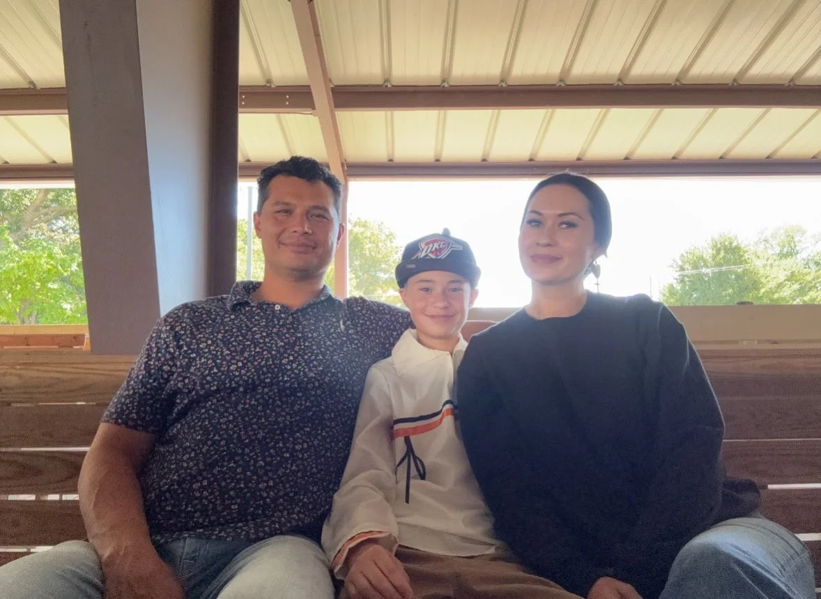 A family of three sitting on a wooden bench indoors near a window with greenery outside. The father is on the left, wearing a black patterned shirt, the son is in the middle wearing a white jacket and a black cap, and the mother is on the right wearing a black sweater.