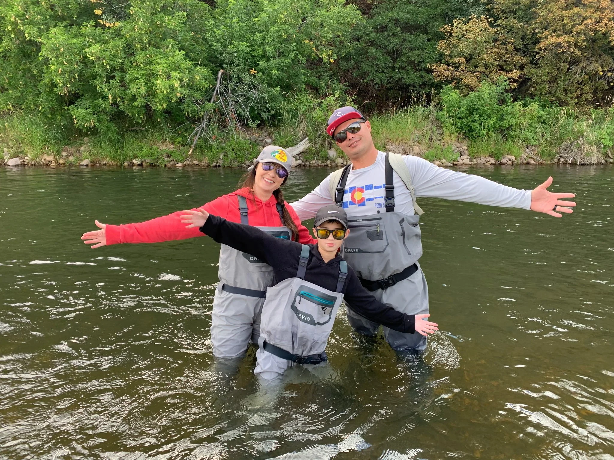 Family of three standing in a river with arms spread wide, wearing fishing gear and sunglasses, with green trees in the background.