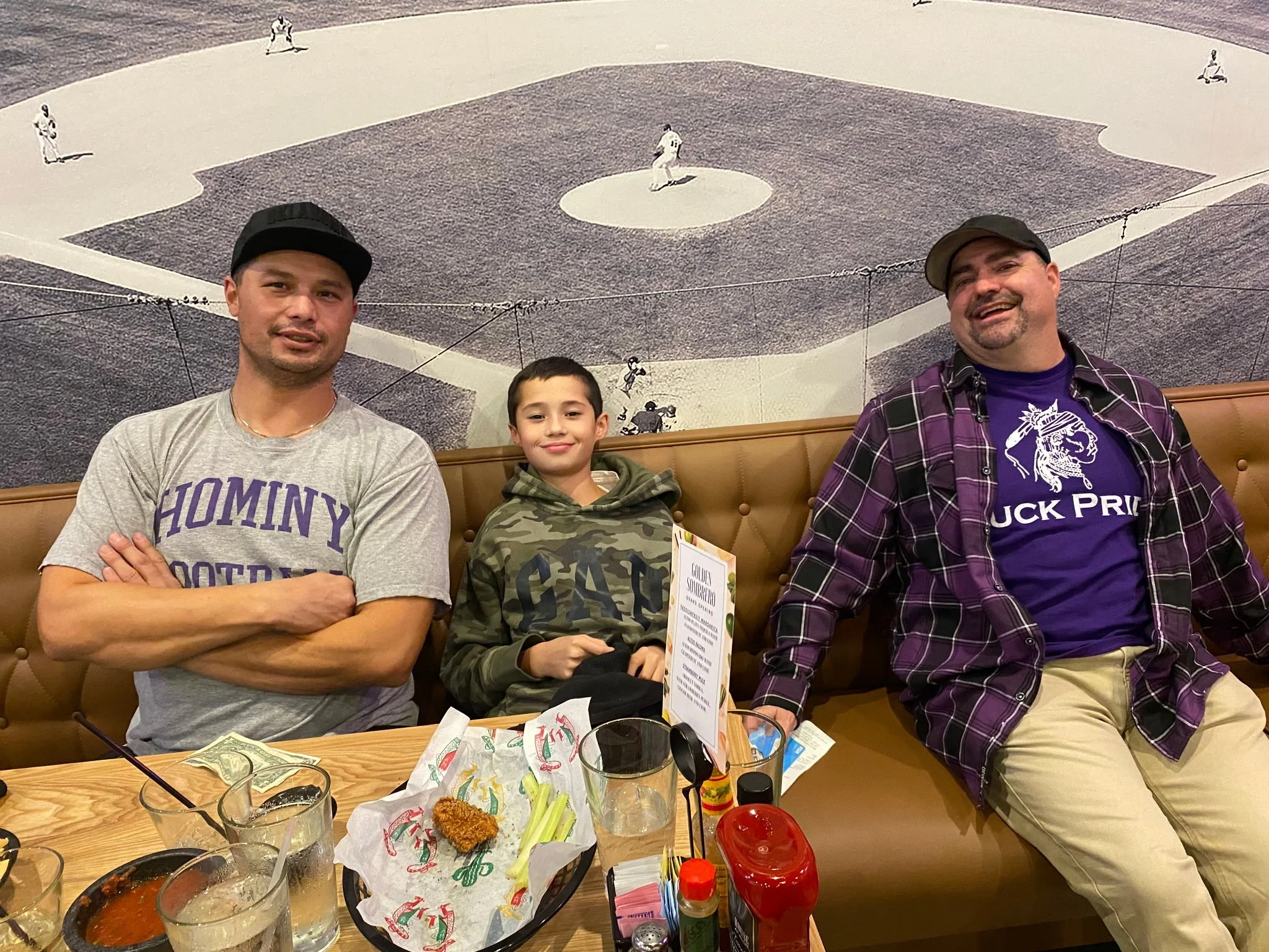 Three people sitting on a booth bench at a restaurant, with a mural of a baseball game on the wall behind them. The person on the left is an adult male in a gray shirt with purple lettering, wearing a black cap. The person in the middle is a young boy wearing a camouflage hoodie with a smile, holding a menu. The person on the right is an adult male in a black cap, purple T-shirt, and a purple plaid shirt unbuttoned over it, smiling. In front of them are drinks, a basket of fried food, and condiments.