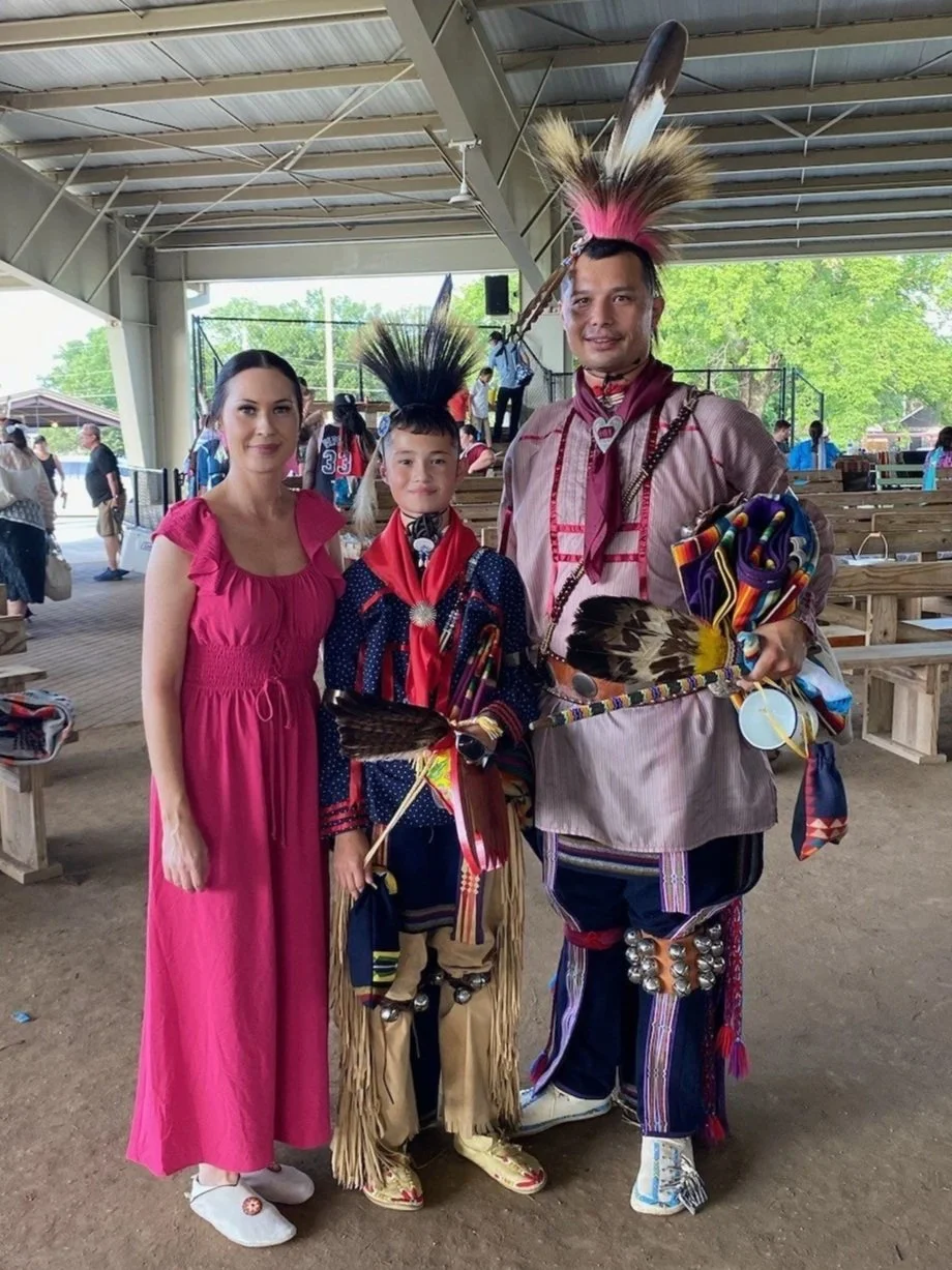 A woman in a pink dress standing next to two people dressed in traditional Native American attire, including feathered headdresses and accessories, inside a covered outdoor pavilion.