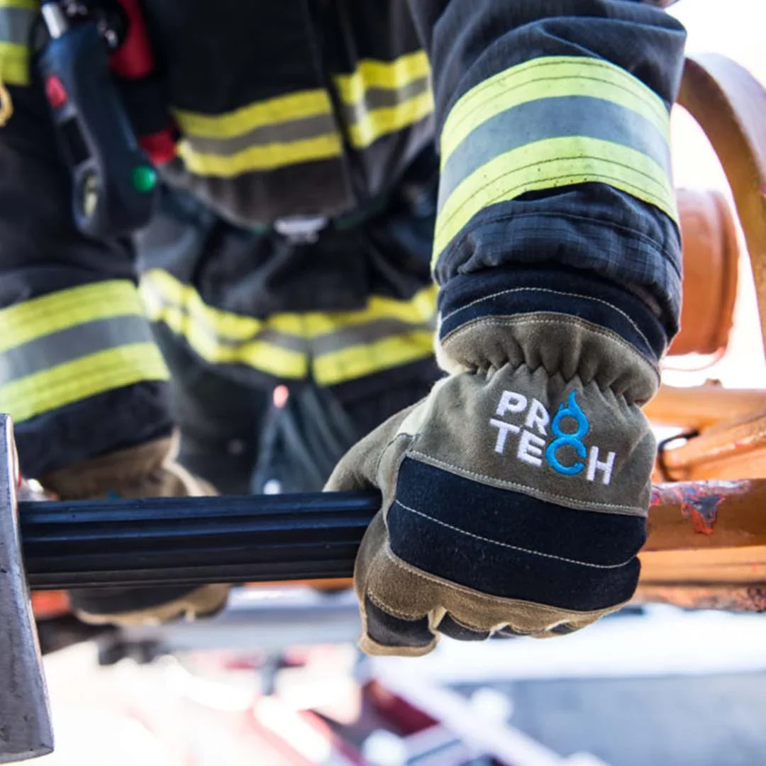 Mano con guante gris que dice Protech en letras blancas con símbolo de llama azul, en un entorno de incendio o rescate, con un uniforme de bombero con rayas amarillas y negras.