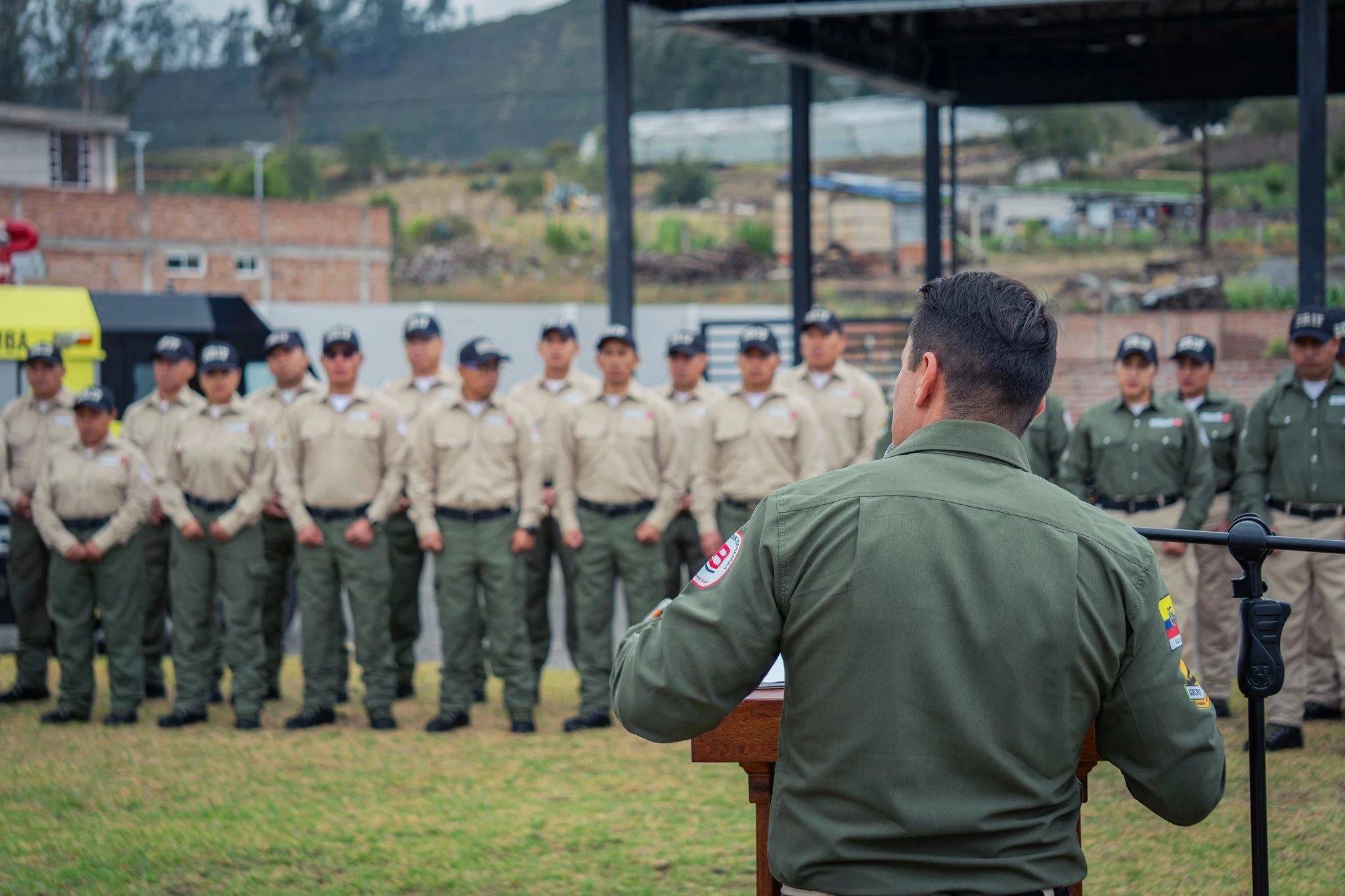 Un grupo de policías uniformados de pie en formación, frente a un oficial que habla en un podio al aire libre, con fondo de área rural y montañas.
