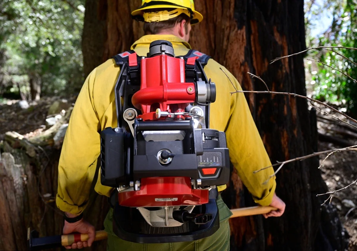 Un bombero con equipo de protección y mochila de agua, usando una herramienta para cortar un árbol en un bosque.