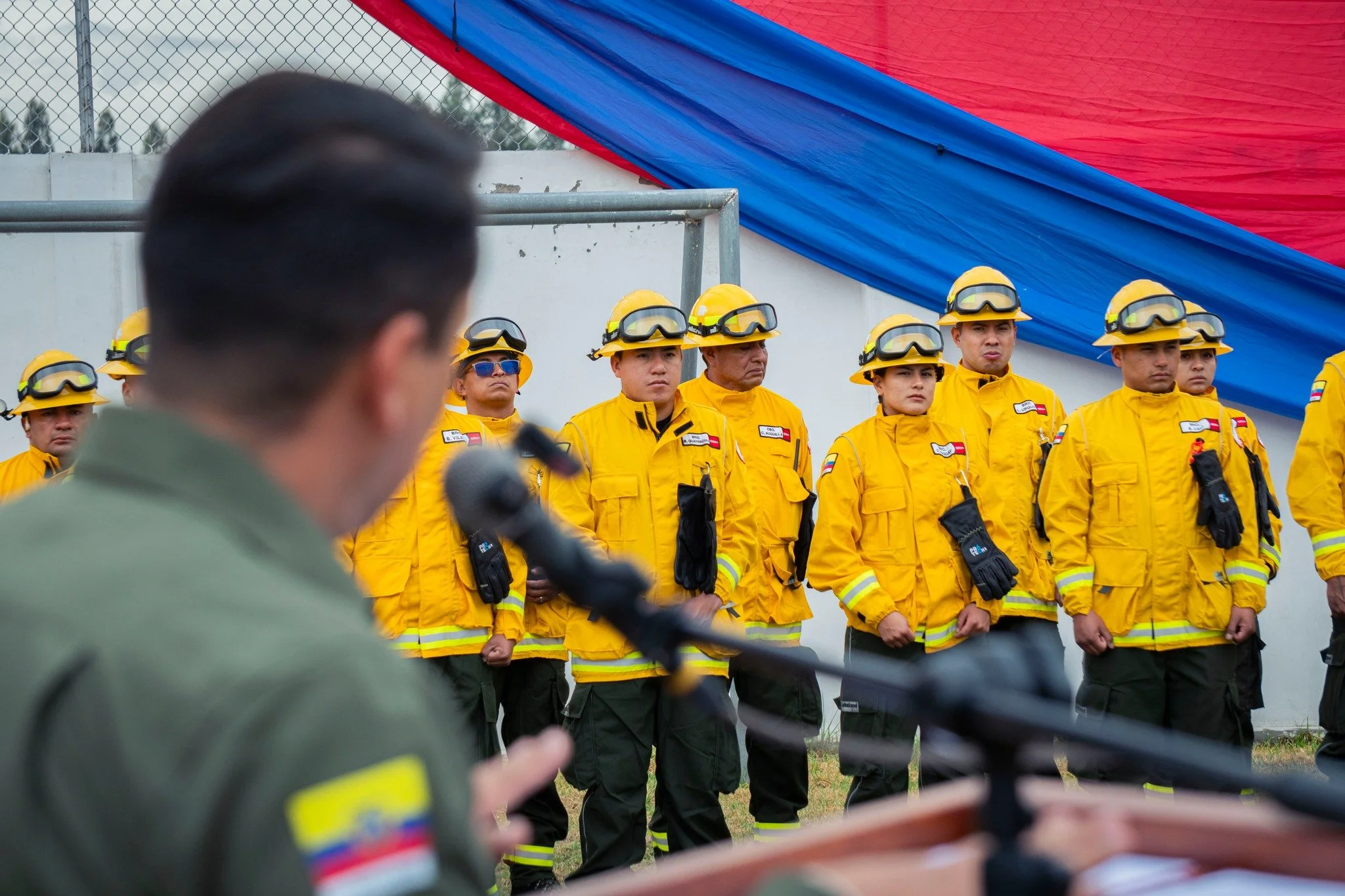 Grupo de bomberos uniformados con casco y ropa de protección durante una ceremonia o instrucción, con una estructura de lona en colores rojo y azul en el fondo.