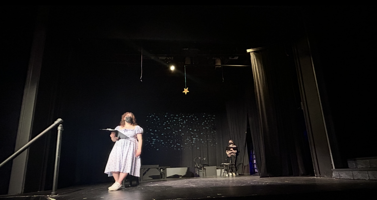 A girl wearing a dress and face mask standing on stage with a script or book in her hand, in a theater with dark curtains, a star decoration hanging from the ceiling, and two chairs on the stage.