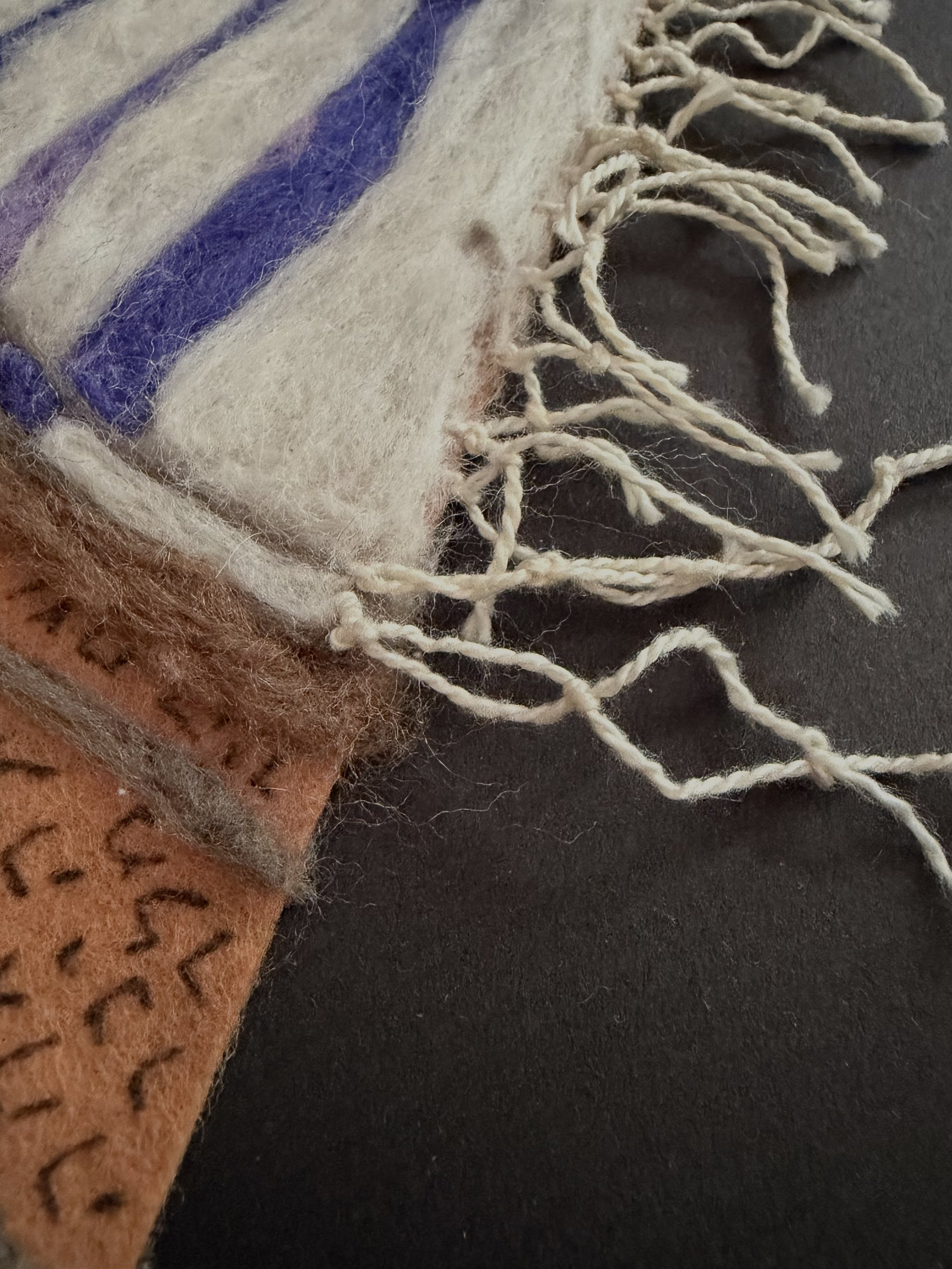 Close-up of a folded, striped Tallis with fringed edges next to a brown textured fabric on a black surface.