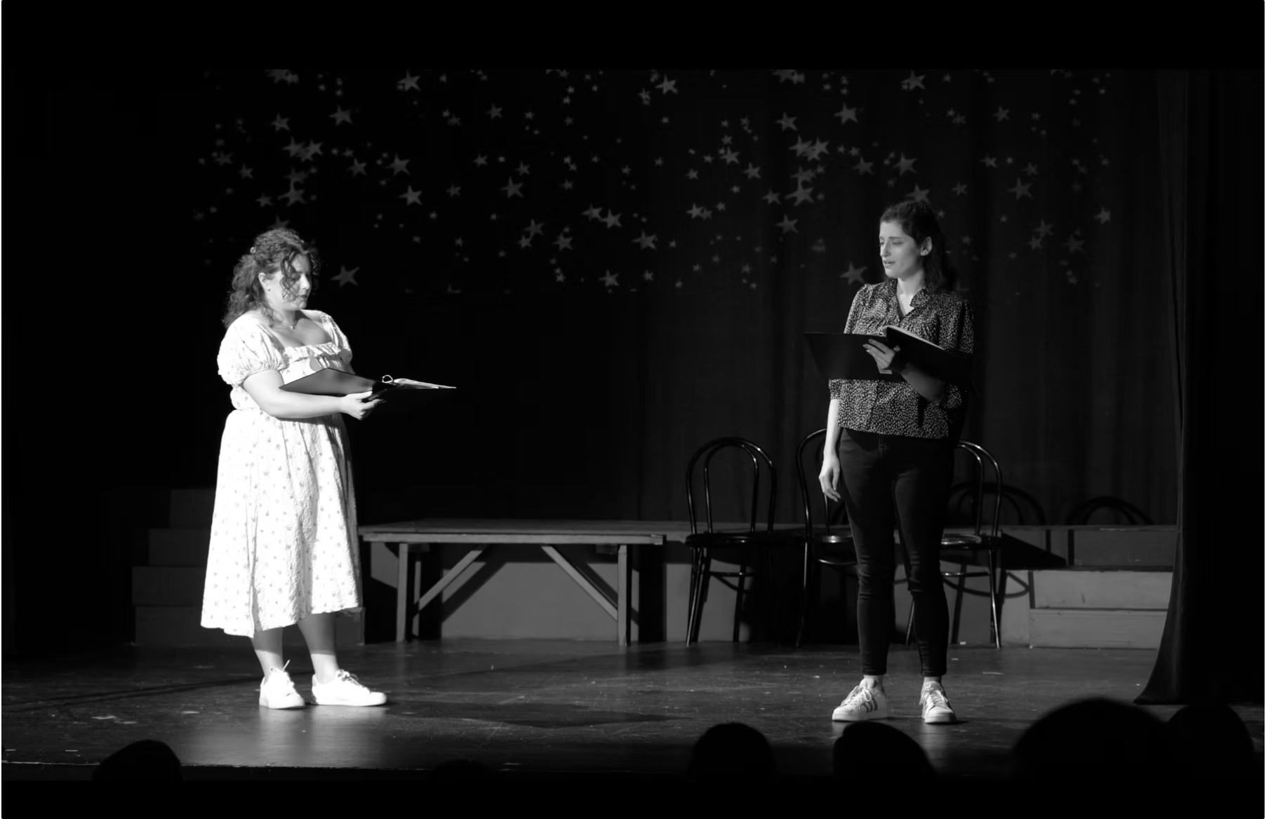 Two women performing on stage, holding scripts, with a backdrop decorated with stars, in a black-and-white photo.