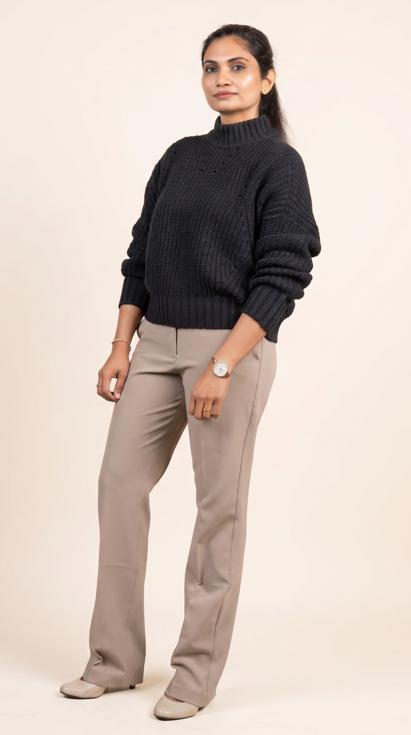 A woman with dark hair styled in a ponytail, wearing a black knitted turtleneck sweater and beige trousers, standing against a plain light-colored background.