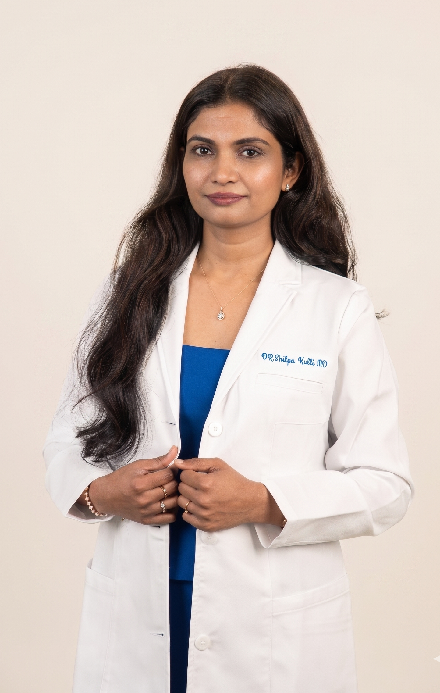 Professional woman in a white medical coat with embroidered name, standing against a plain background, holding her coat with both hands.