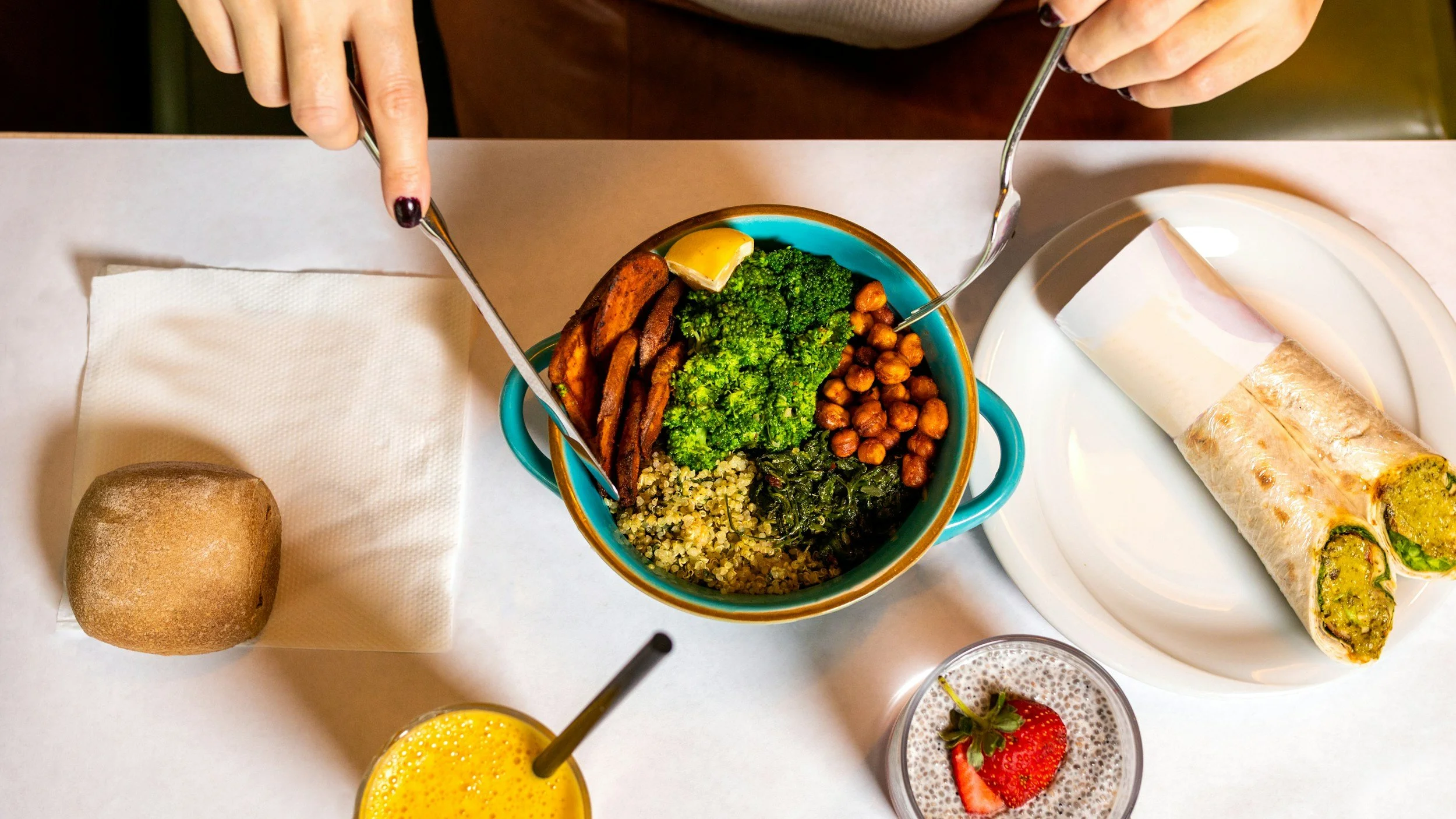 A colorful meal with a bowl of mixed vegetables and grains, a slice of lemon, a roll bread, a spring roll with green filling, a strawberry, and a glass of orange juice on a white table.