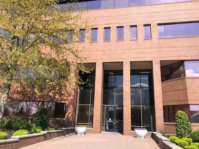 Modern brick office building with large glass doors and windows, surrounded by landscaped bushes and trees under a blue sky.