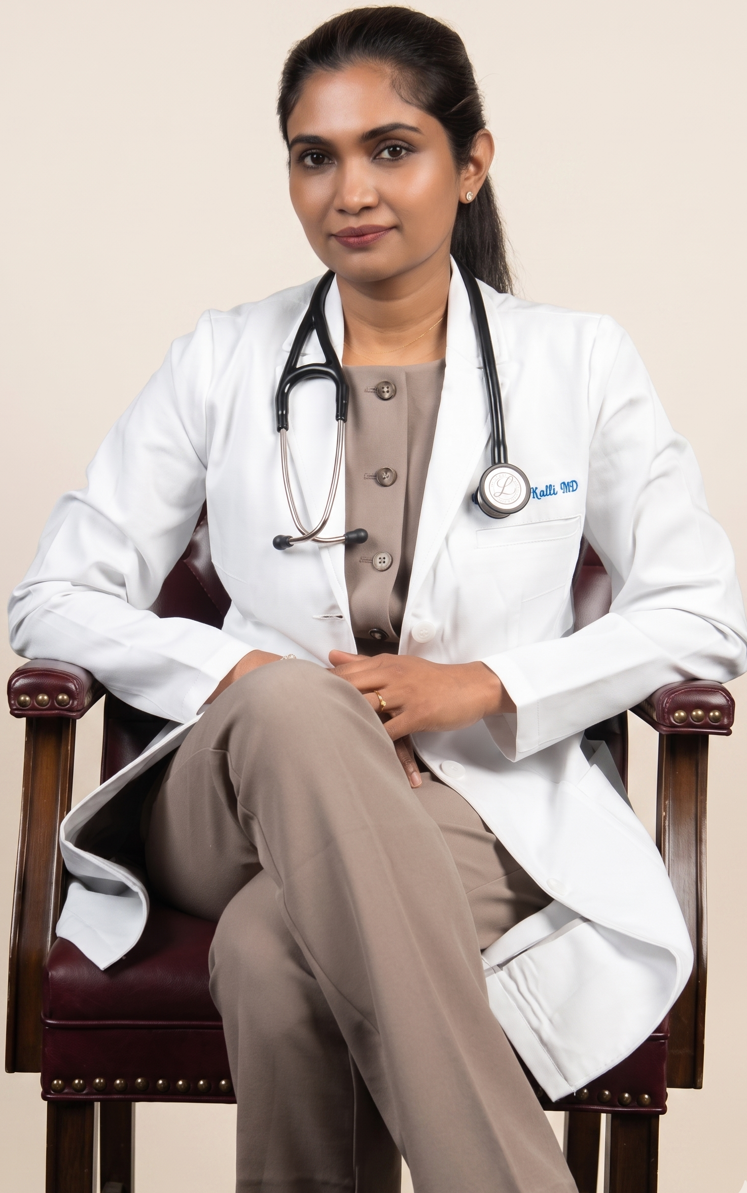 A female doctor sitting confidently in a chair, wearing a white lab coat with a stethoscope around her neck, and a beige outfit underneath, against a plain background.