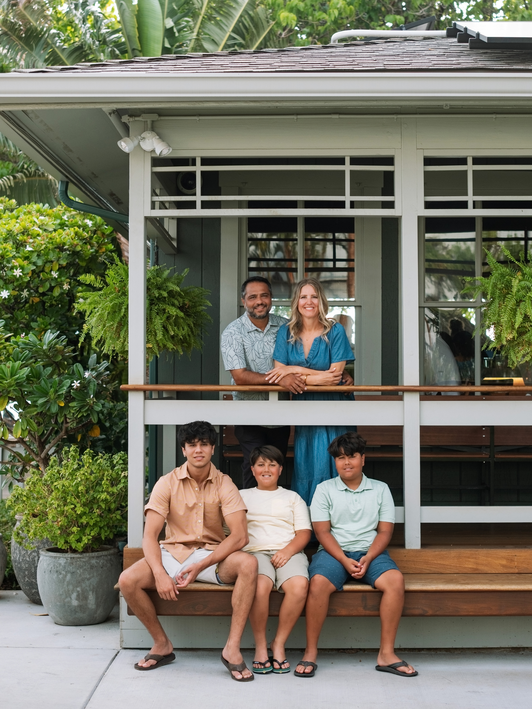 A diverse group of five people, three children and two adults, sitting and standing on the steps of a porch surrounded by greenery, smiling at the camera.