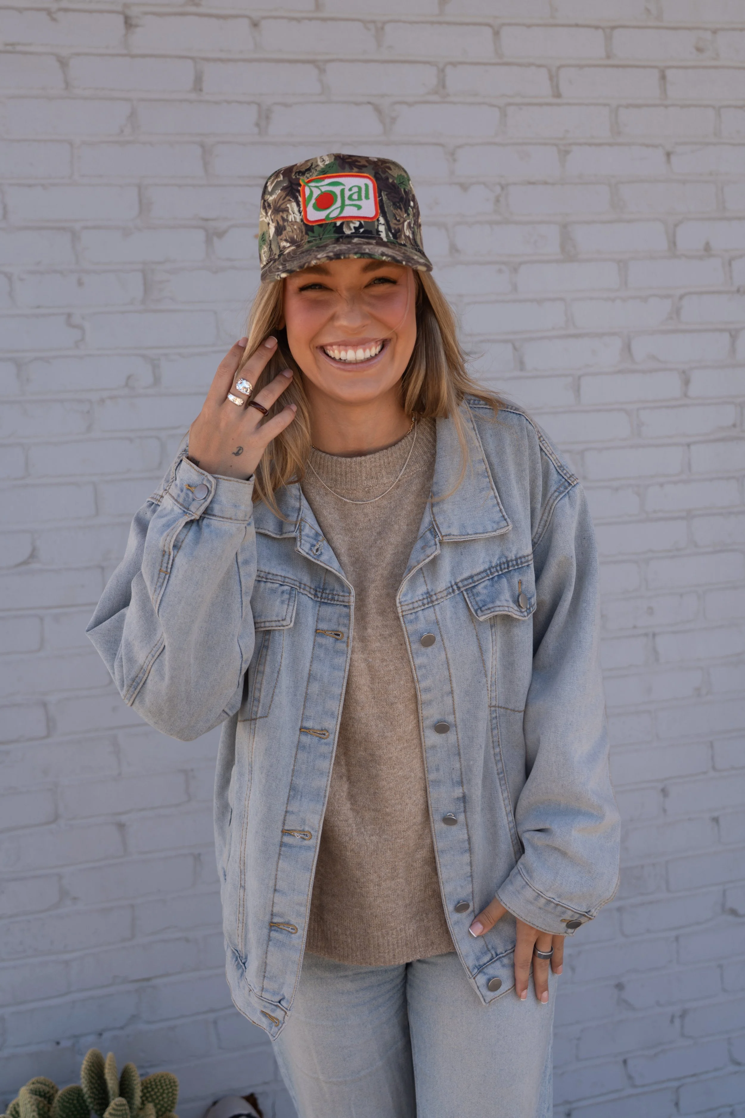 A woman smiling and talking on her phone, wearing a camouflage baseball cap with a patch that says 'jal' and a marijuana leaf, a light denim jacket, and light-colored jeans, standing against a white brick wall.