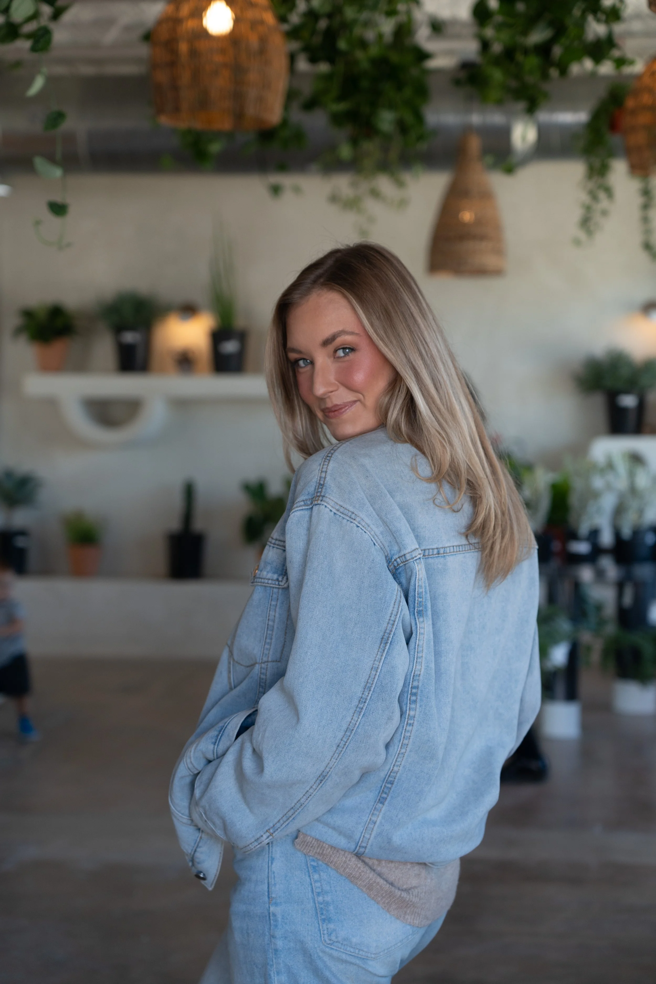 A young woman with blonde hair, wearing a denim jacket and jeans, turns back and smiles in an indoor space decorated with hanging rattan lamps and potted plants.