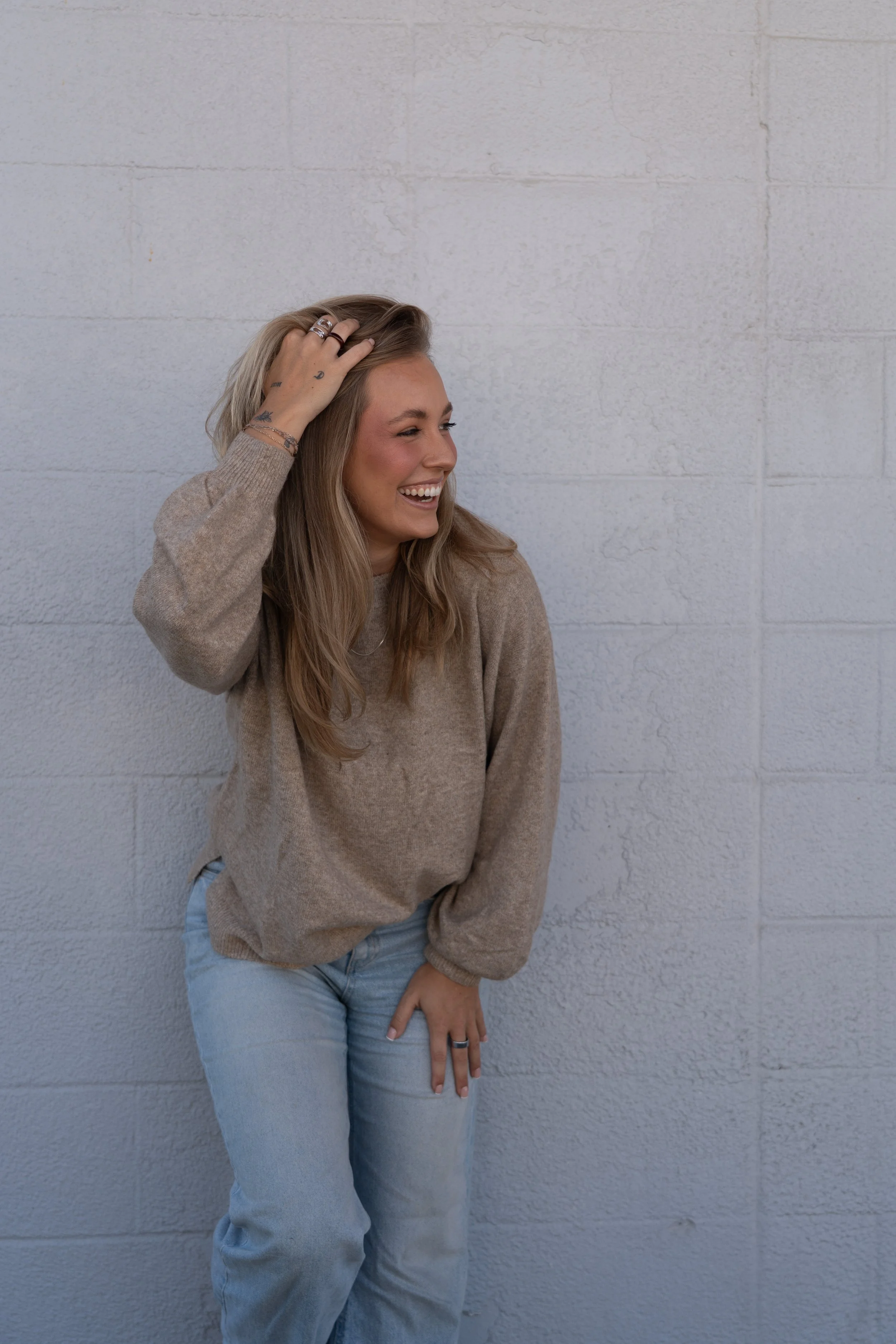 A young woman with long blonde hair laughing and smiling, wearing a beige sweater and light blue jeans, standing against a plain white brick wall.