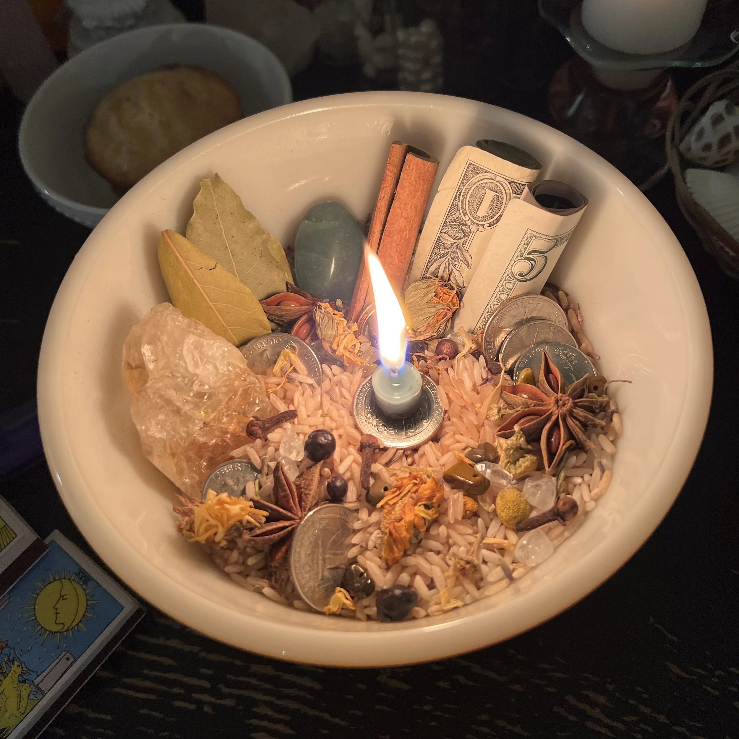 A white bowl filled with various herbs, spices, and money, with a lit candle in the center.