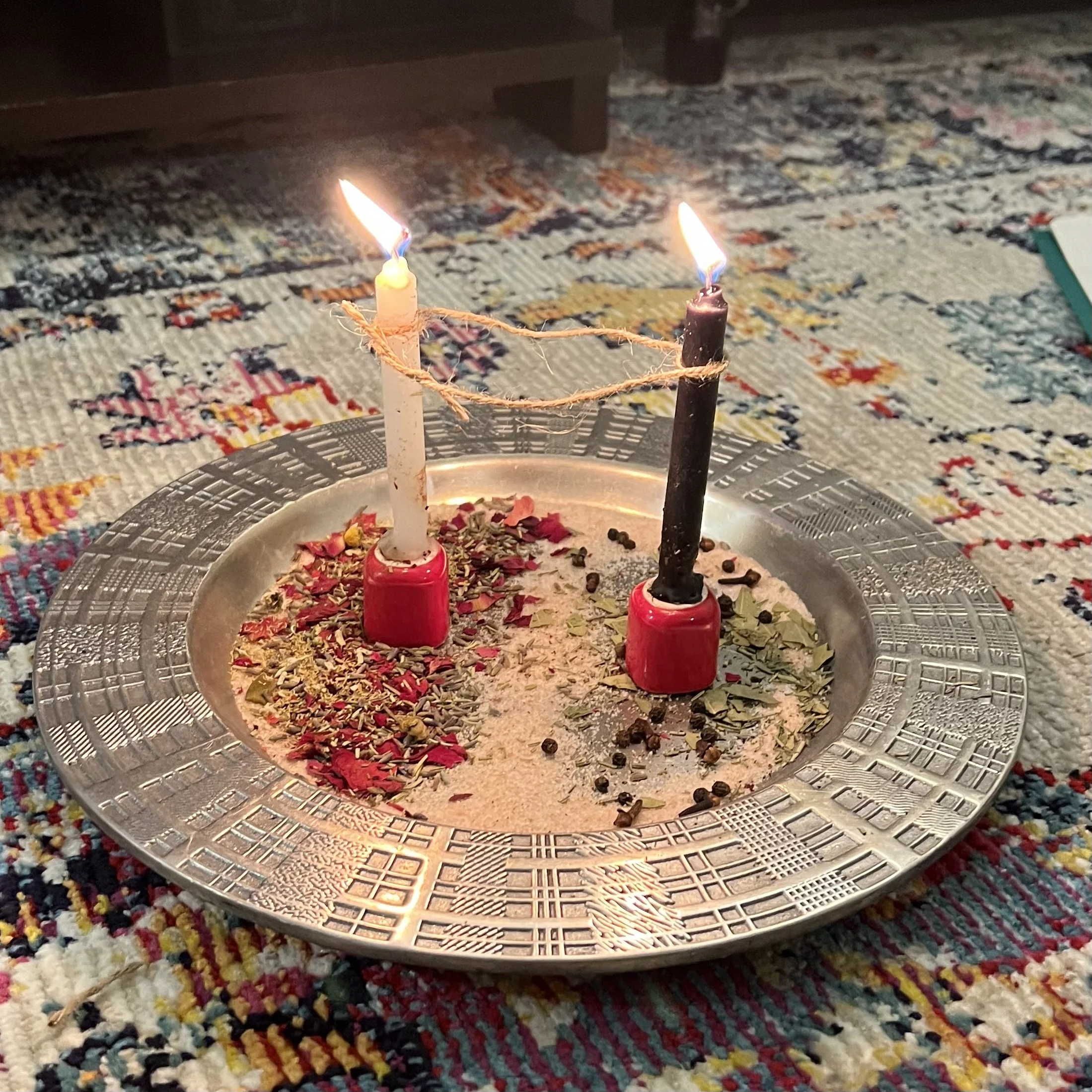 Diya lamp with two candles, one still lit, and a lit matchstick, on a decorative metal tray filled with rice, spices, and flower petals, on a colorful woven rug.
