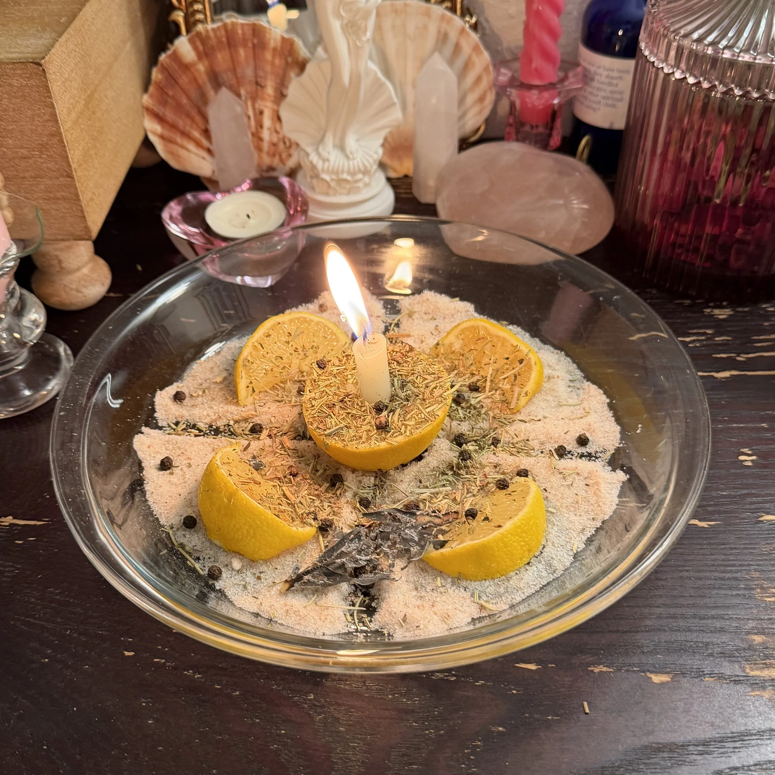 Decorative glass bowl with dried orange slices, sage, and black peppercorns, topped by a lit candle at the center.