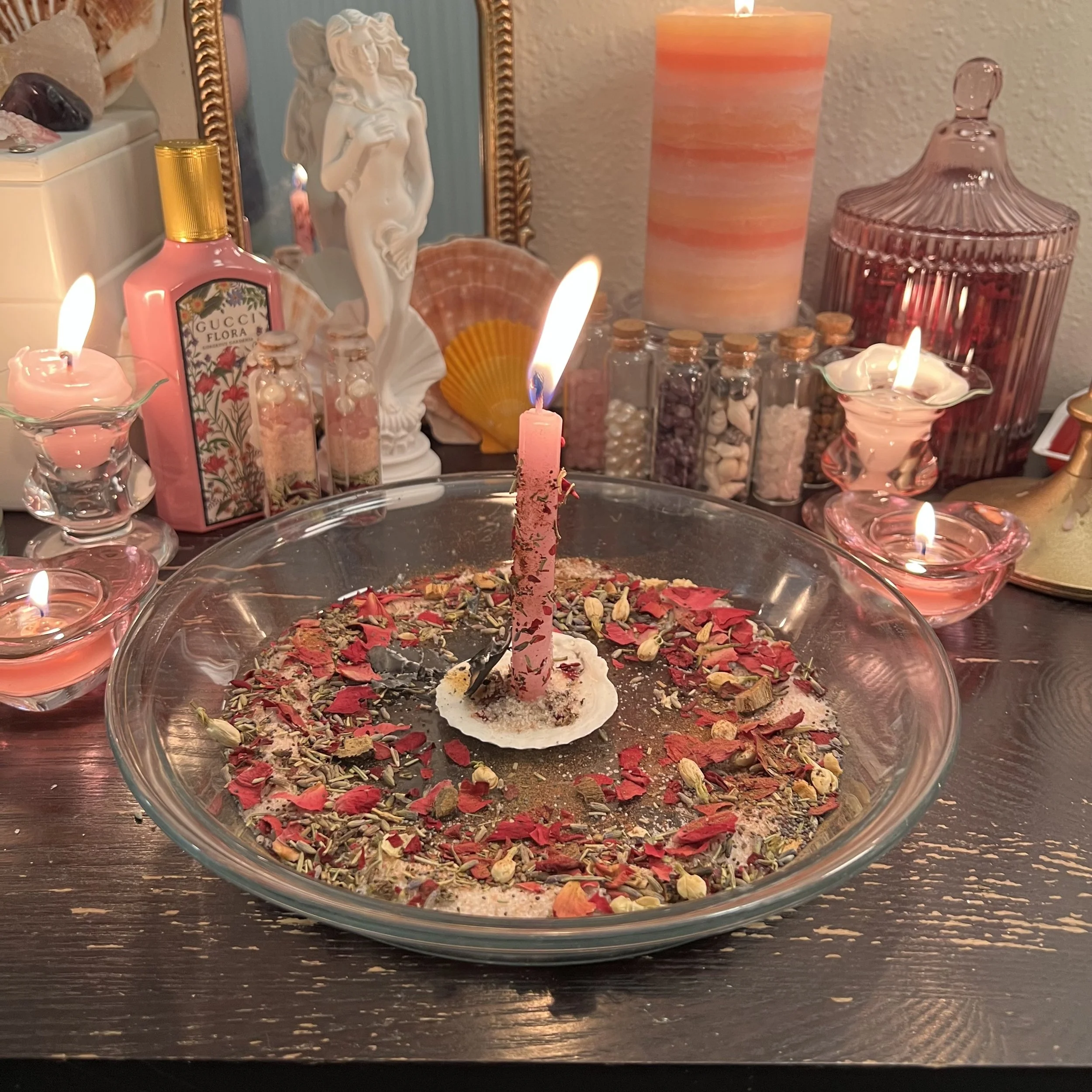 A table decorated with candles, dried flower petals, and various decorative bottles and figurines, including a mother-of-pearl shell, a statue, and a pink perfume bottle, with a lit birthday candle in the center.