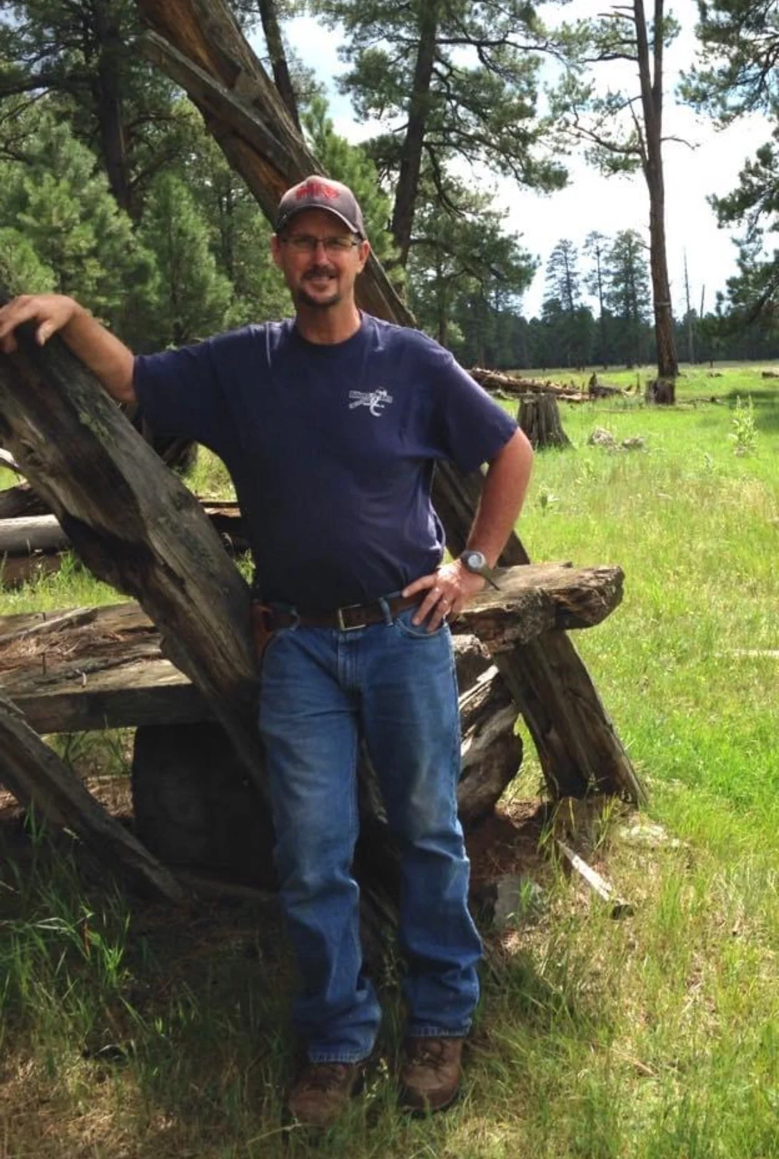A man standing outdoors with a fallen tree in the background, smiling and wearing a cap, glasses, a dark blue t-shirt, jeans, and boots.