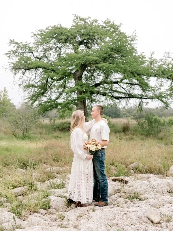 A couple standing on rocks outdoors, embracing each other with a woman holding a bouquet of flowers, large green tree in the background.