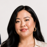 A woman with long black hair, wearing gold hoop earrings and makeup, smiling slightly against a light gray background.