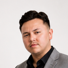 Portrait of a young man with short dark hair, wearing a gray blazer and black shirt against a plain white background.