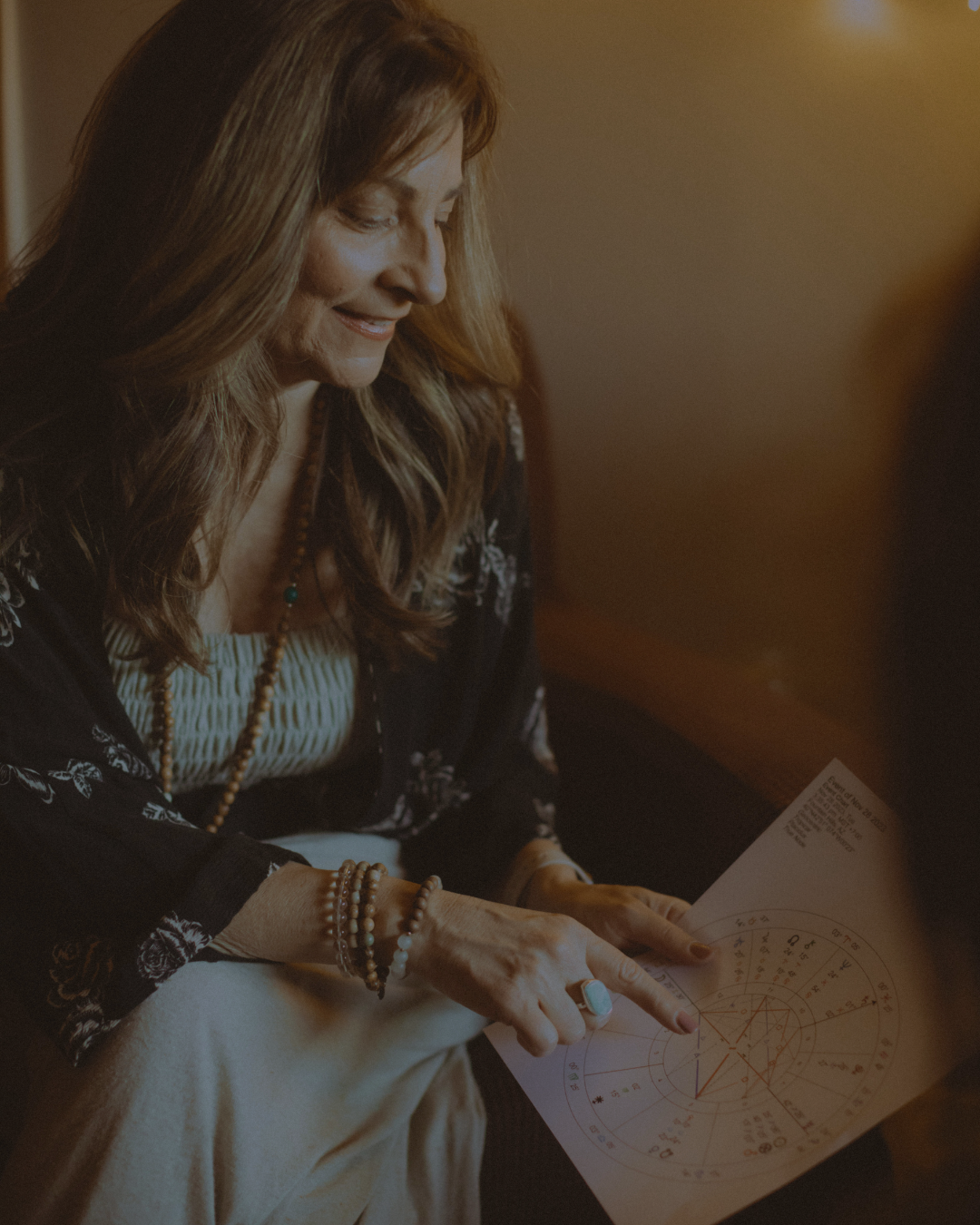 A woman with long brown hair wearing a black floral blouse, light-colored pants, and jewelry, is sitting and reading a paper with astrological charts or diagrams.