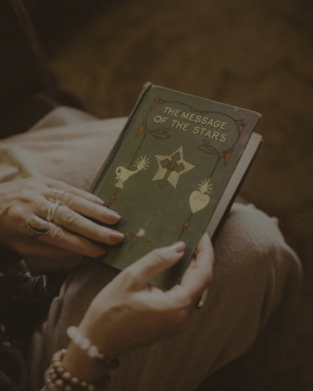 Person holding a book titled 'The Message of the Stars' with decorative illustrations. Only hands and part of the person's legs are visible.