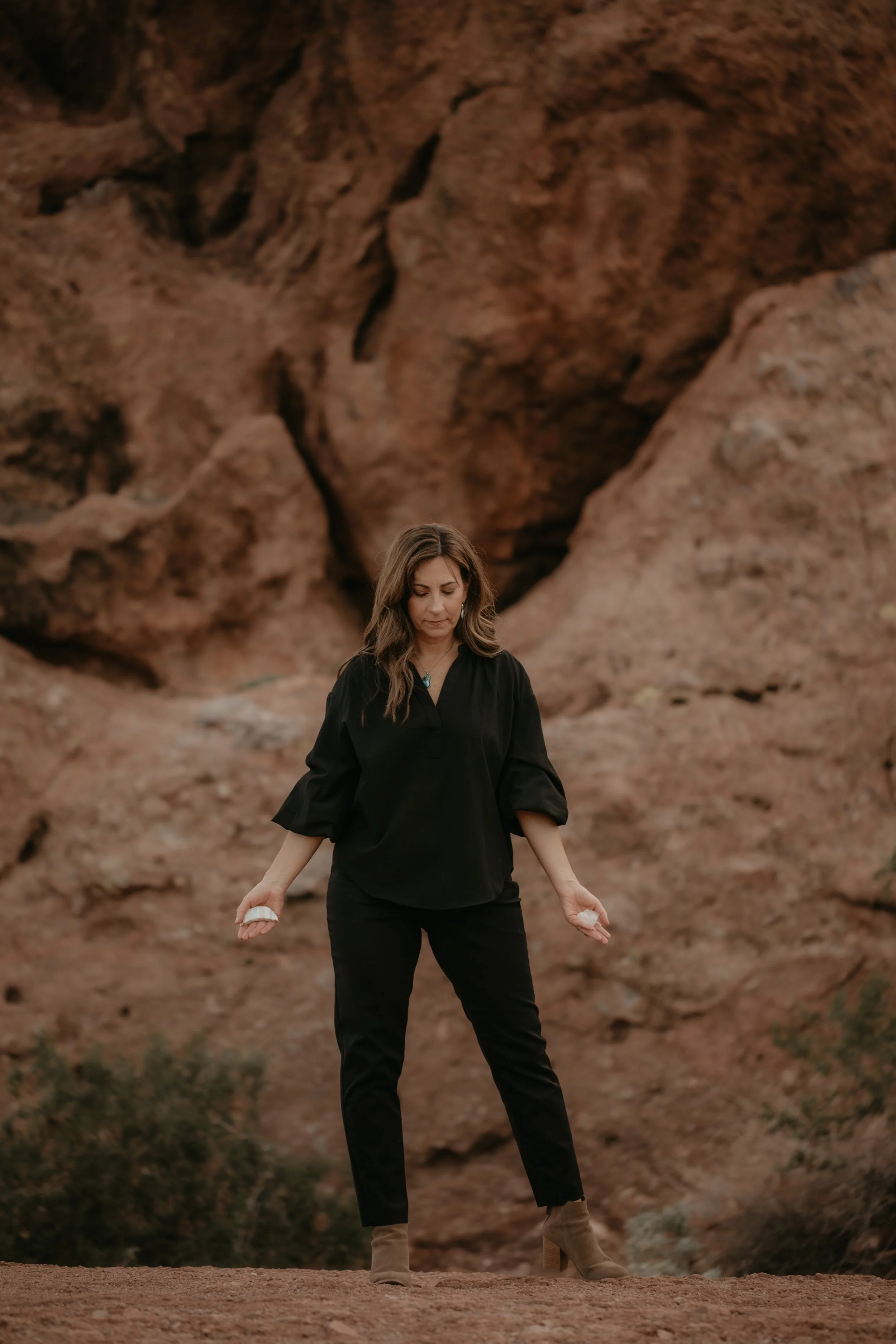 Woman in black outfit standing outdoors in front of large rock formation with reddish-brown hues.