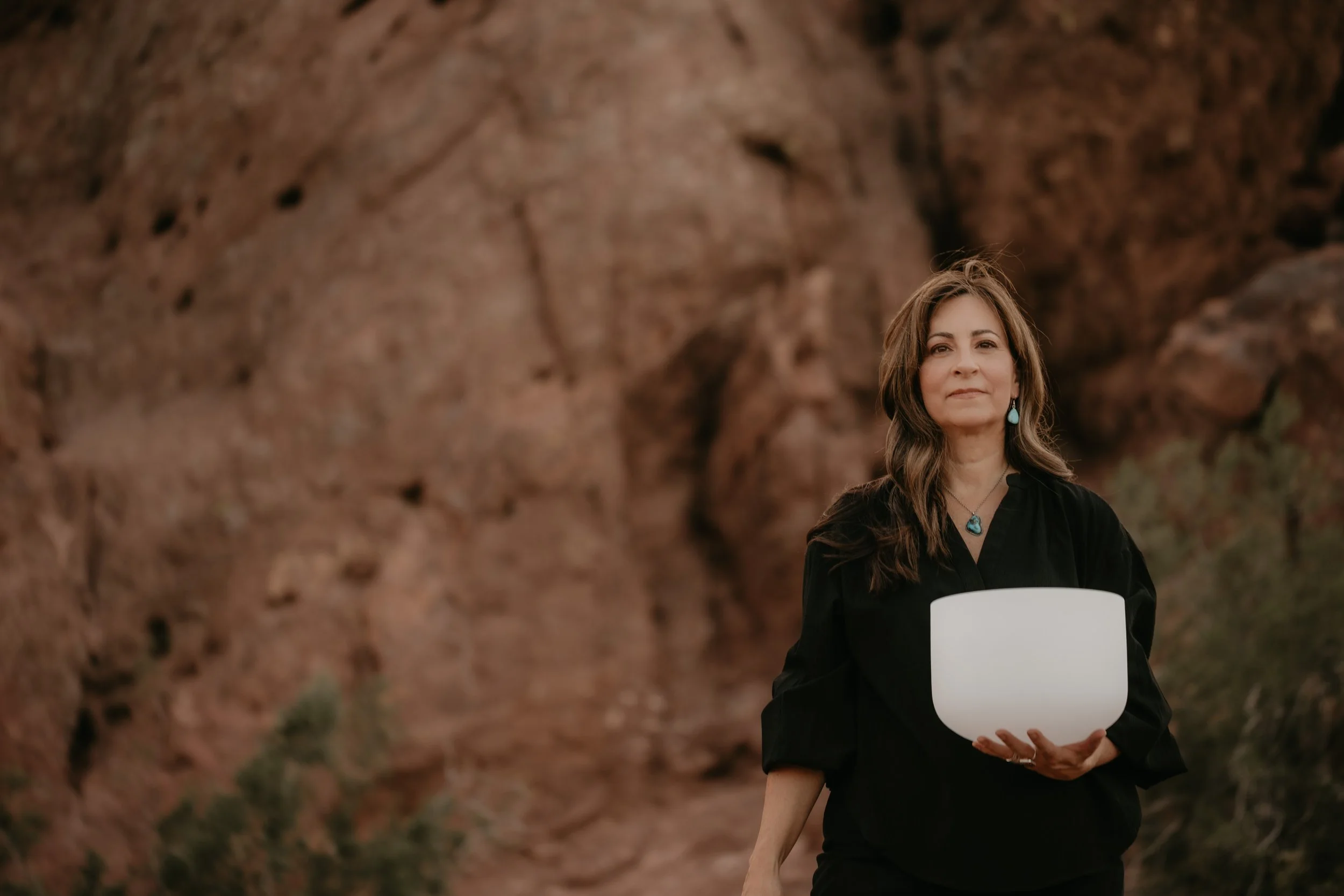 A woman with long brown hair, wearing turquoise jewelry, standing outdoors near large rock formations, holding a large white bowl.