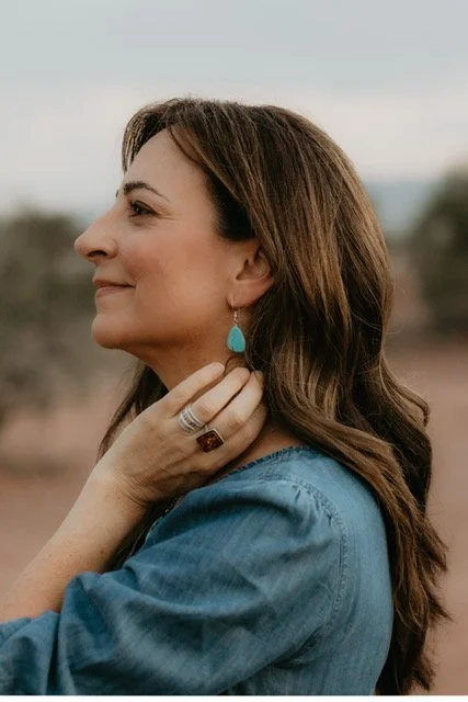 Side profile of a woman with long brown hair wearing a blue denim shirt, turquoise earring, and rings, outdoors during daytime.