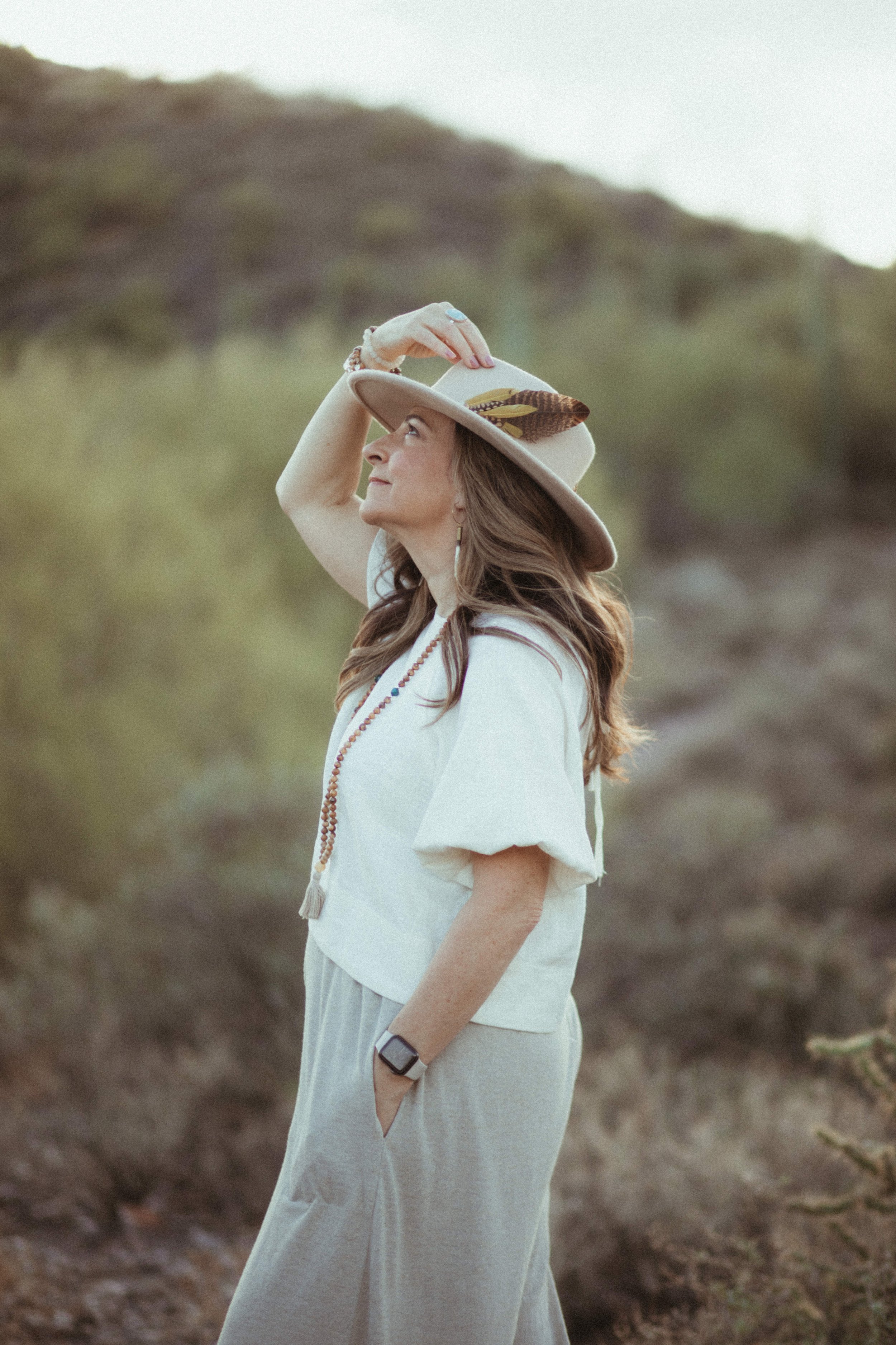 A woman with long brown hair wearing a beige hat decorated with feathers and a white blouse, standing outdoors on a grassy hill, looking upward with her hand on her hat.