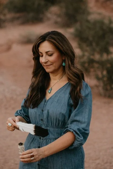 A woman with long brown hair wearing a blue dress and turquoise jewelry, holding a stick of sage.