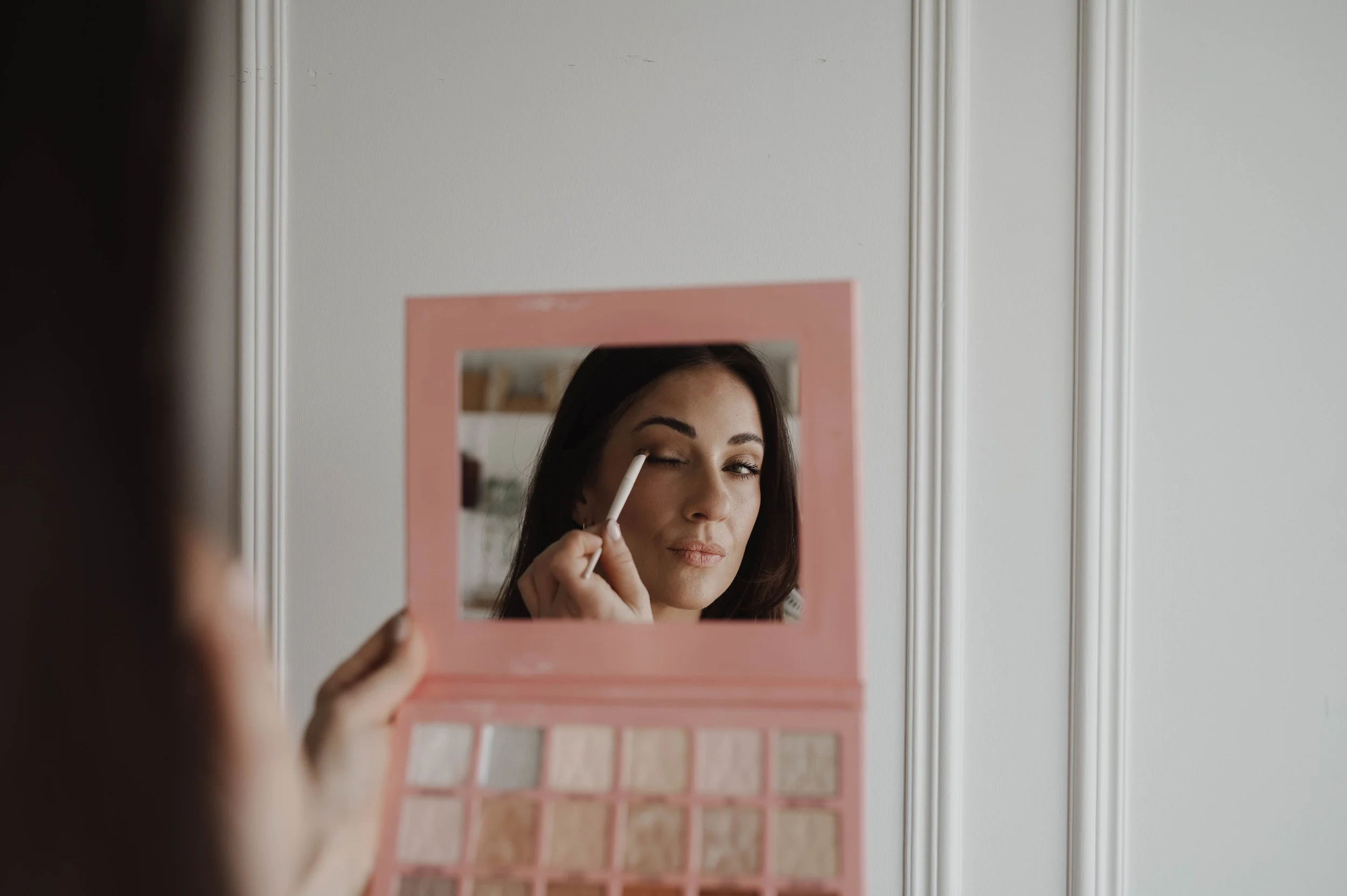 A woman applying makeup while looking into a mirror, seen through a pink makeup palette held in front of the camera.