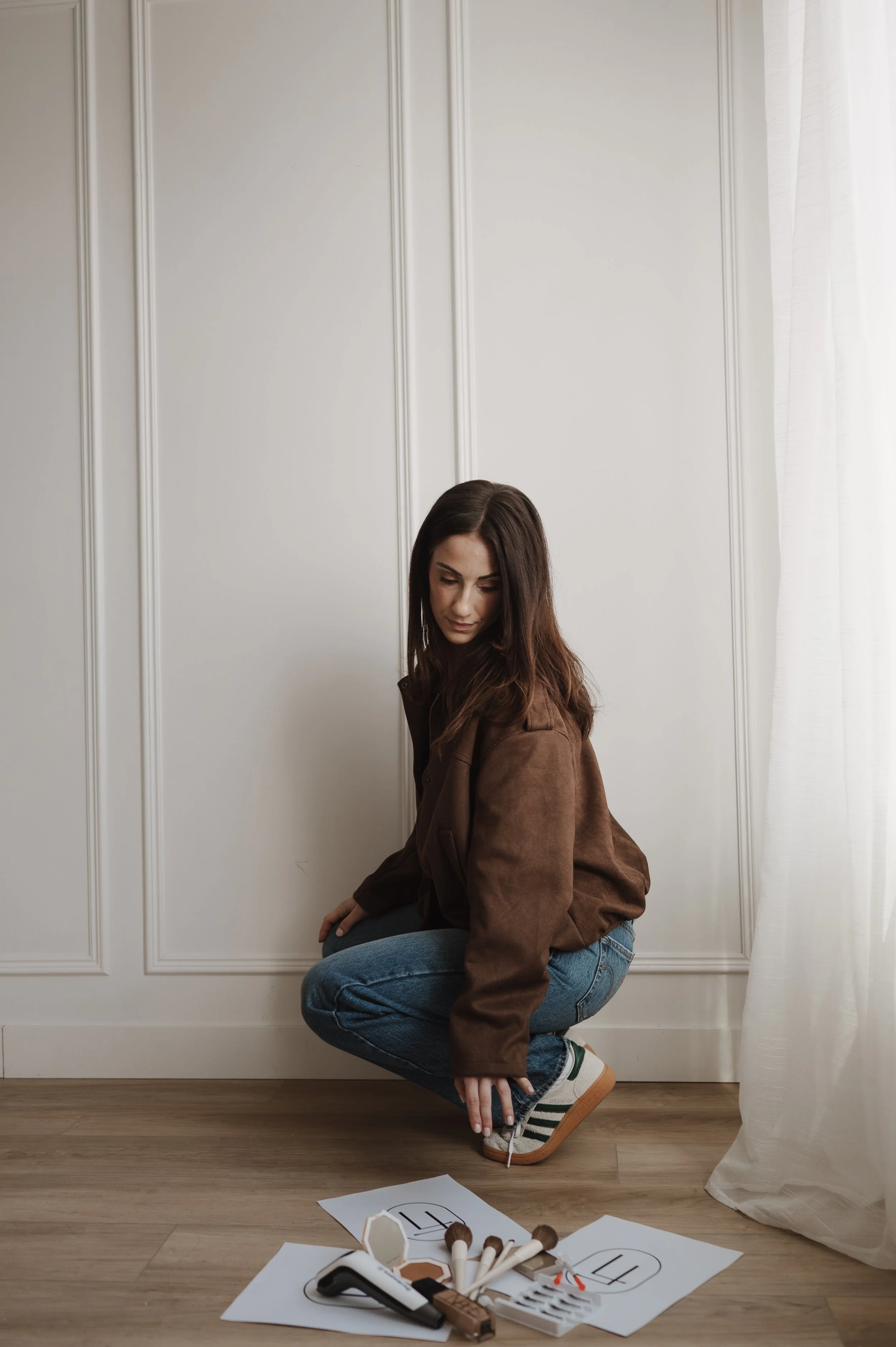 A woman in casual clothes crouches on a wooden floor, with papers, makeup items, and makeup guides scattered in front of her. She is indoors near a white wall with vertical paneling and white curtains.