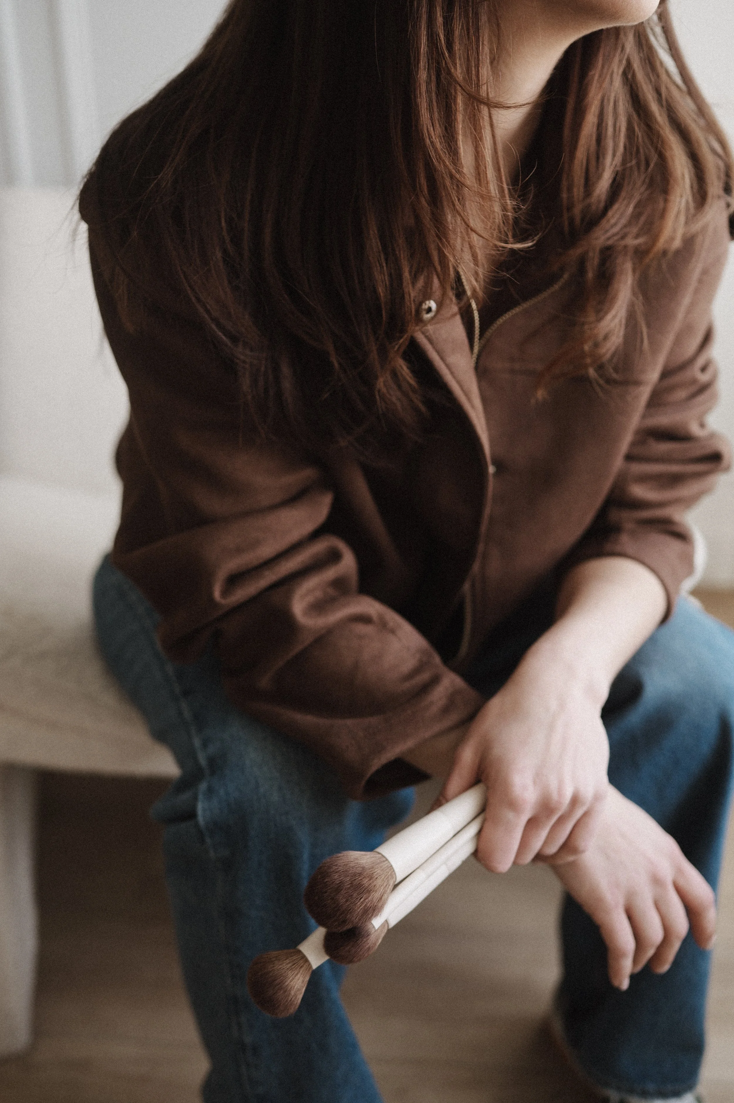 A person dressed in a brown jacket and blue jeans is sitting and holding a white makeup brush with brown bristles.