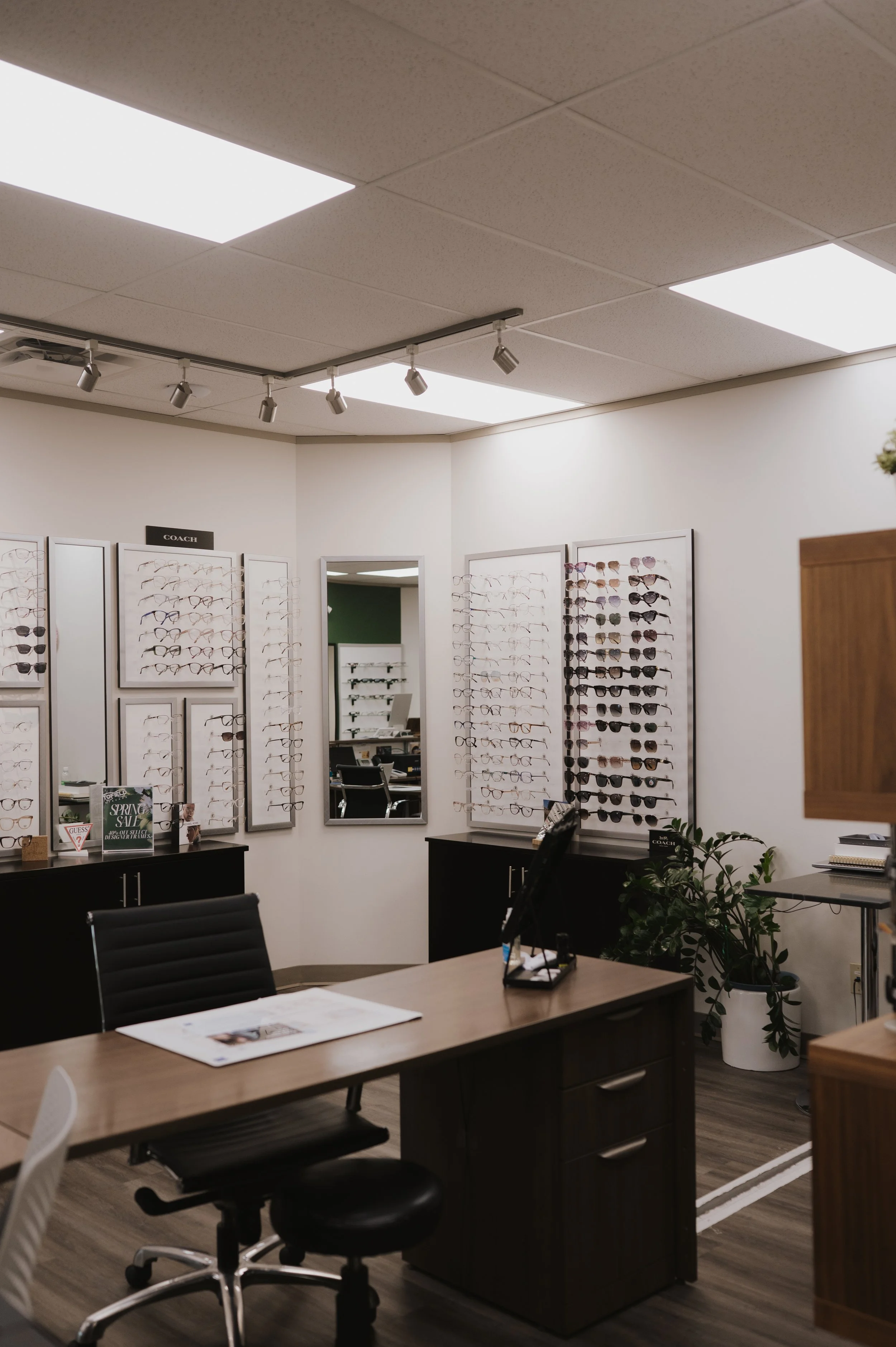 Inside a sunglasses store with display racks of various sunglasses, a desk with papers and office supplies, and office chairs. The ceiling has fluorescent lights and a track lighting system.