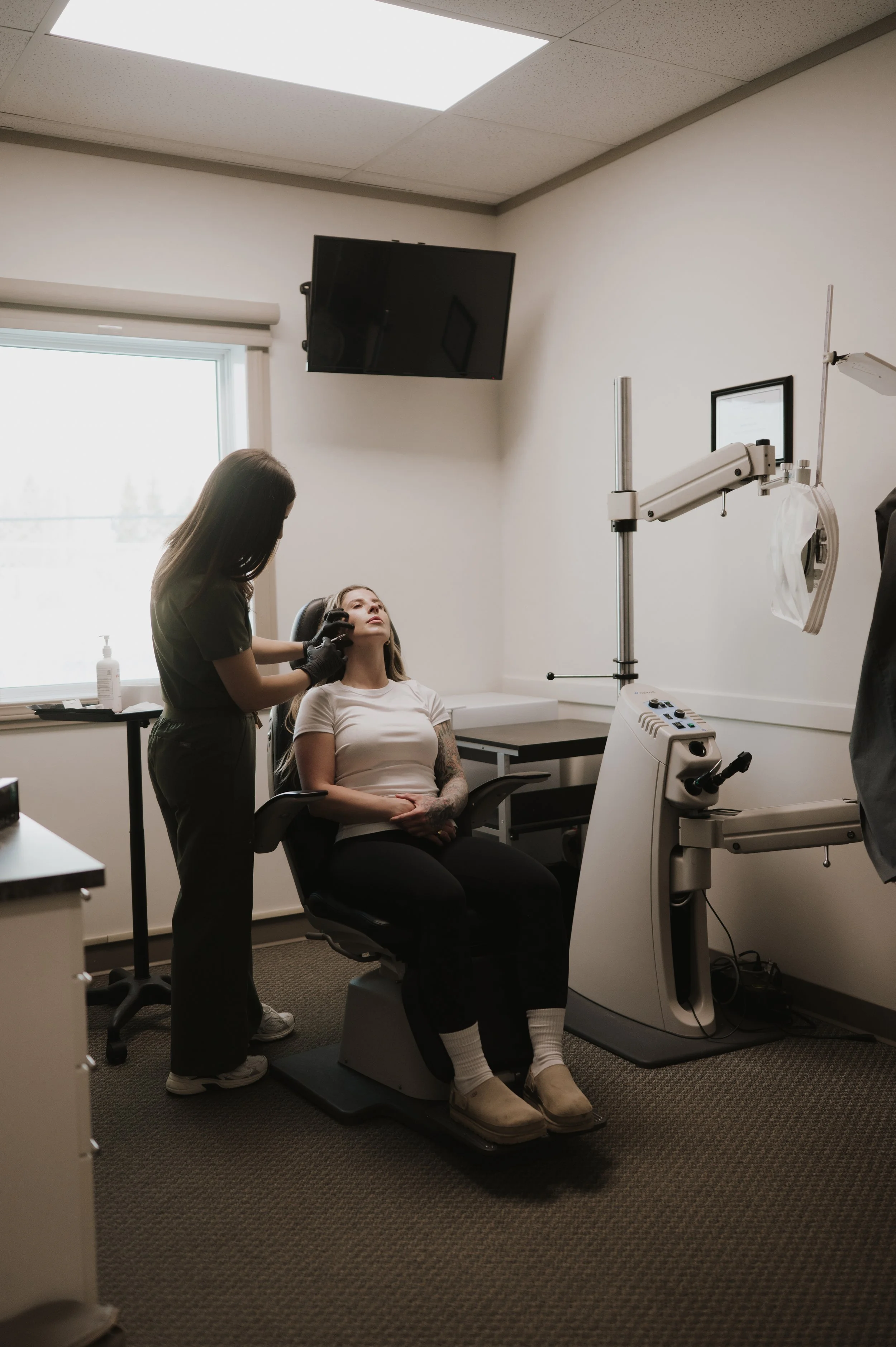 A woman receives a cosmetic or dermatological treatment from a healthcare professional in a medical office. The woman is seated in a specialized chair, while the healthcare worker stands next to her, wearing gloves and administering the procedure. Equipment and a monitor are visible in the room.