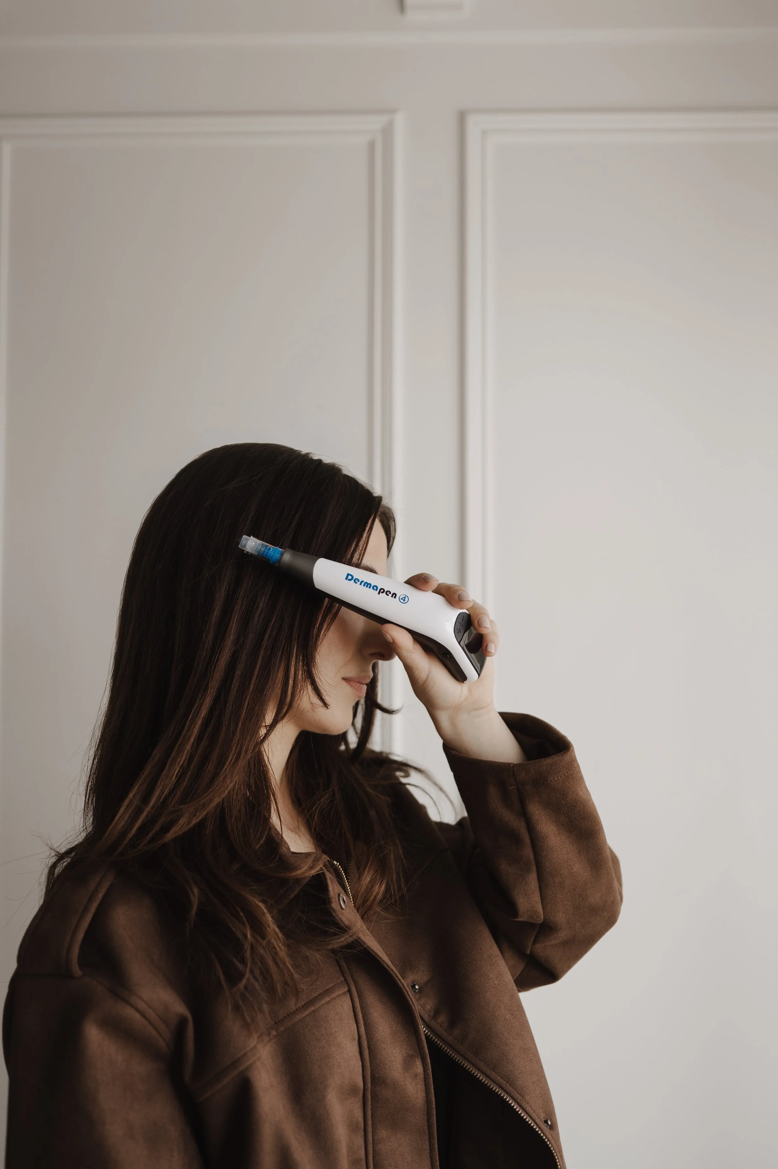 A woman with long brown hair is holding a DermaPen device to her forehead, wearing a brown jacket, in front of a plain white wall.