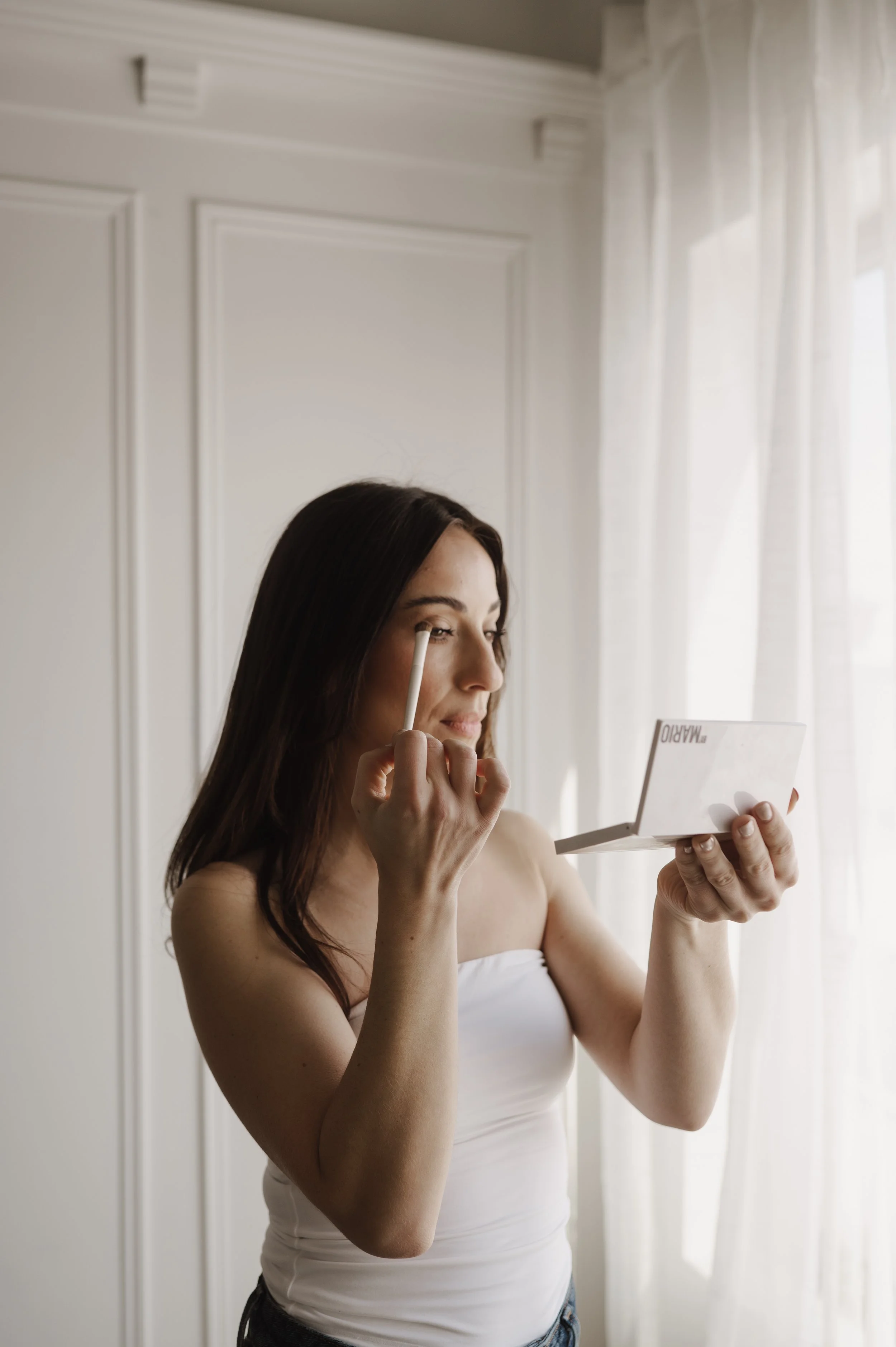 A woman with long dark hair applying makeup with an eyeshadow brush while looking into a mirror, standing in a room with white walls and curtains.