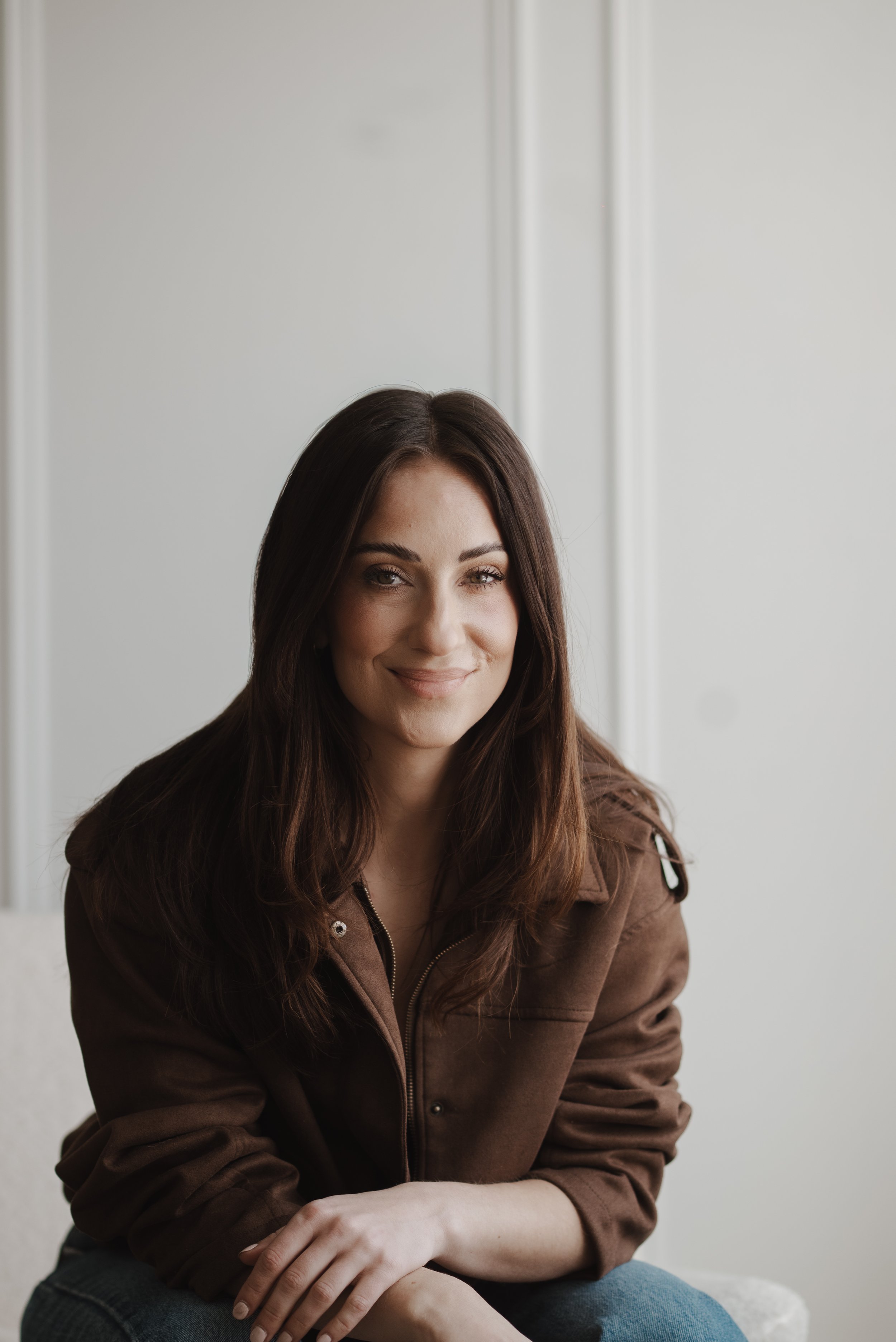 A woman with long brown hair, smiling, wearing a brown jacket, sitting against a plain white wall.