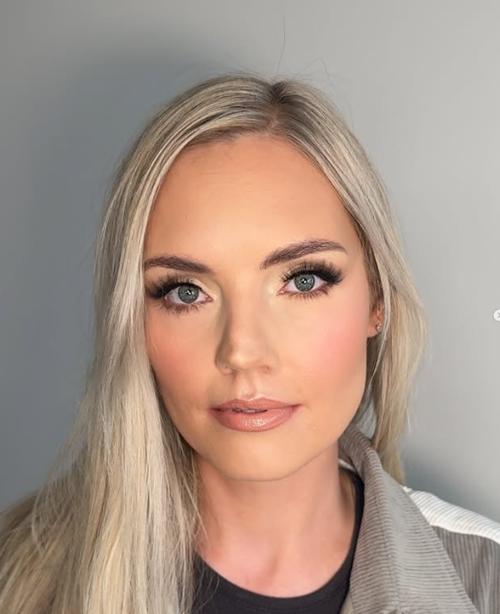 Close-up of a woman with long blond hair and blue eyes, wearing makeup, a light gray jacket, and a black shirt, against a plain gray background.