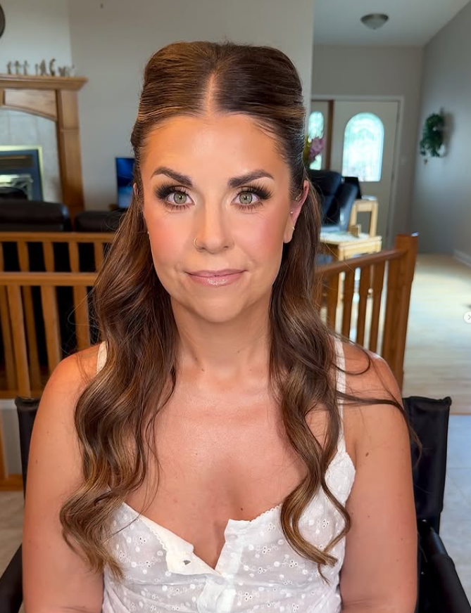 A woman with brown, wavy hair and light skin, wearing a white top with a slight v-neck, sitting indoors in a well-lit room with a wooden staircase and furniture in the background.