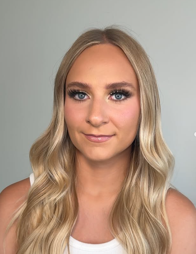 A young woman with long, wavy blonde hair and blue eyes, wearing makeup, a white sleeveless top, and smiling softly against a plain light background.