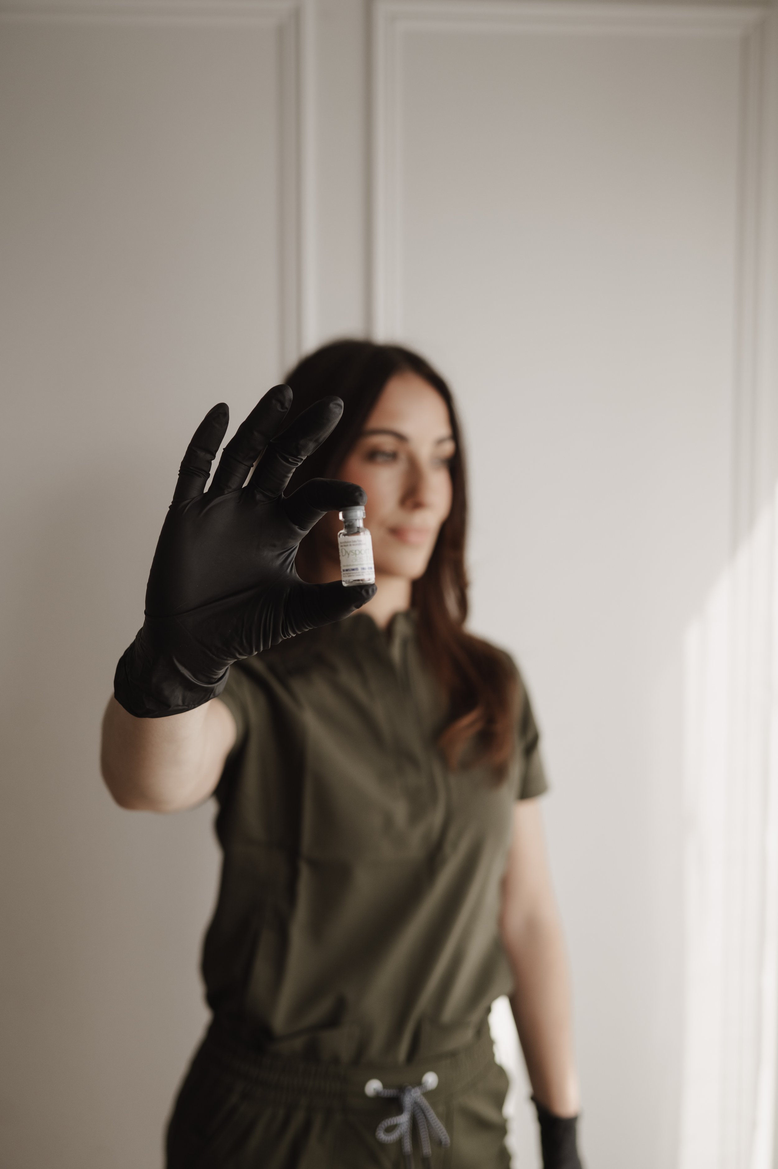 A woman with dark hair wearing a khaki t-shirt and shorts, holding a small vial of vaccine or medication in front of her face with her right hand, which is covered with a black glove, in a neutral indoor setting.