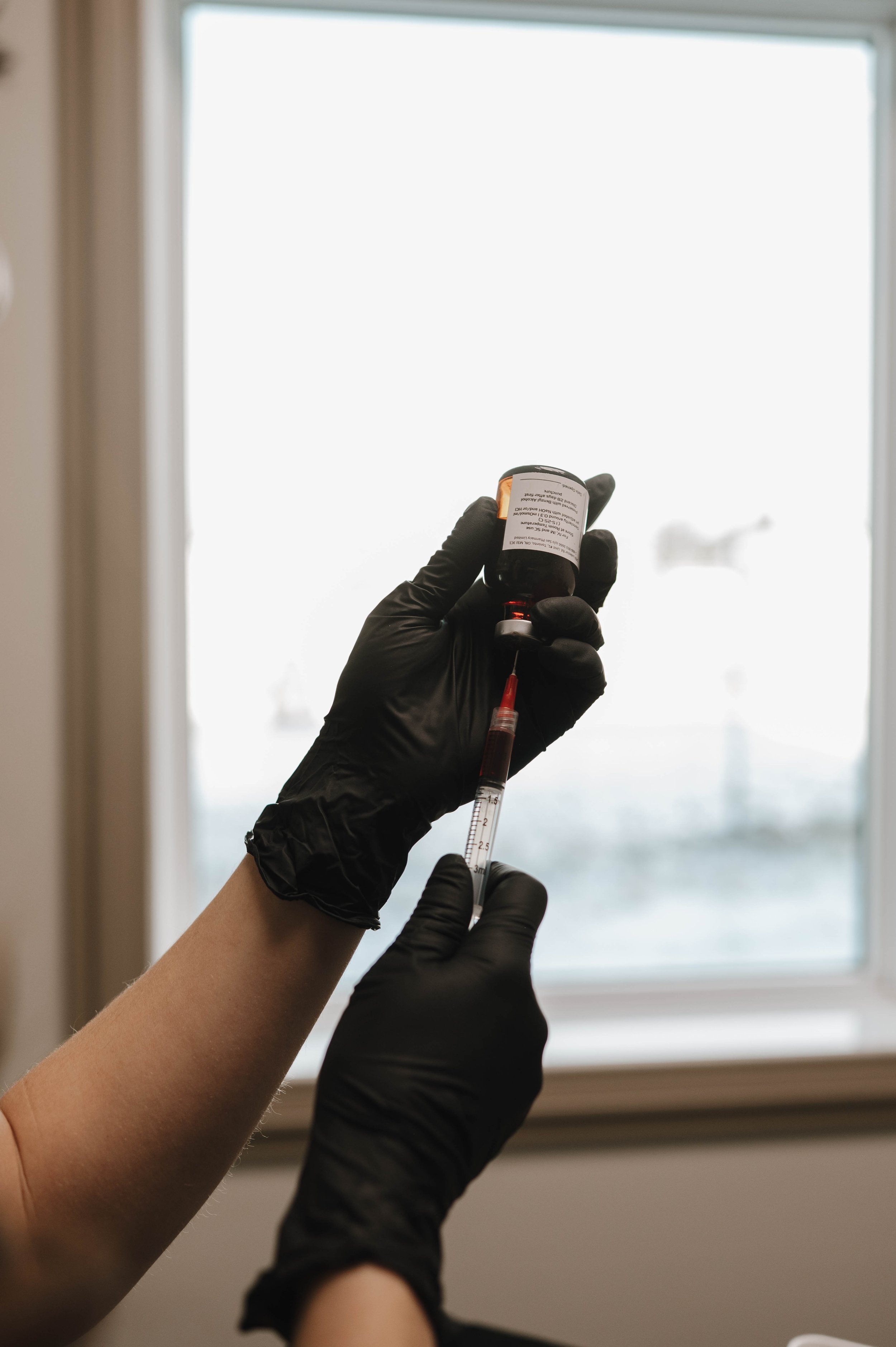 A person wearing black gloves holds a small vial and a syringe in front of a window, with the sky and clouds visible outside.