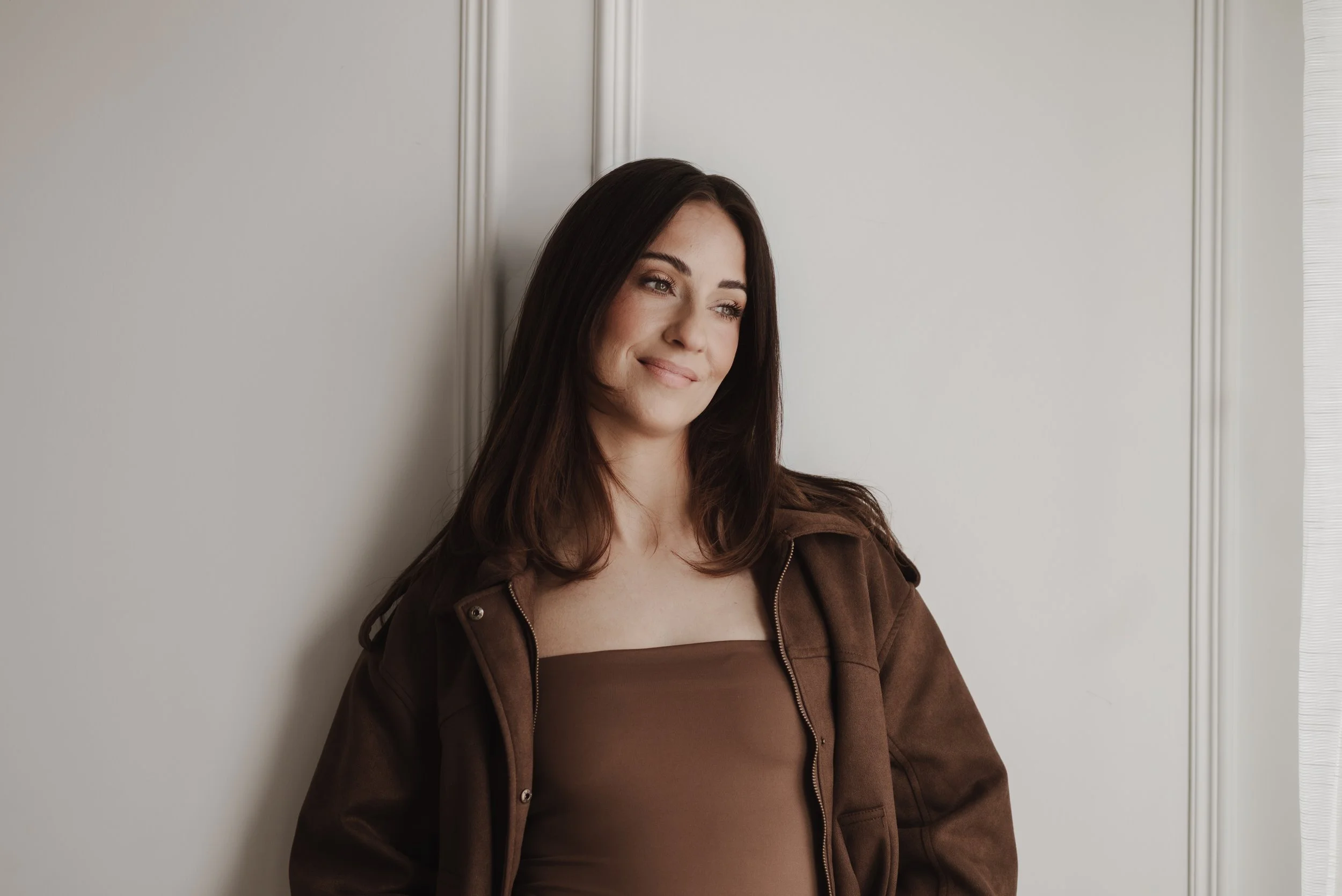 A young woman with long dark hair, wearing a brown jacket and a brown top, smiles softly while leaning against a light-colored wall with decorative molding, near a window.