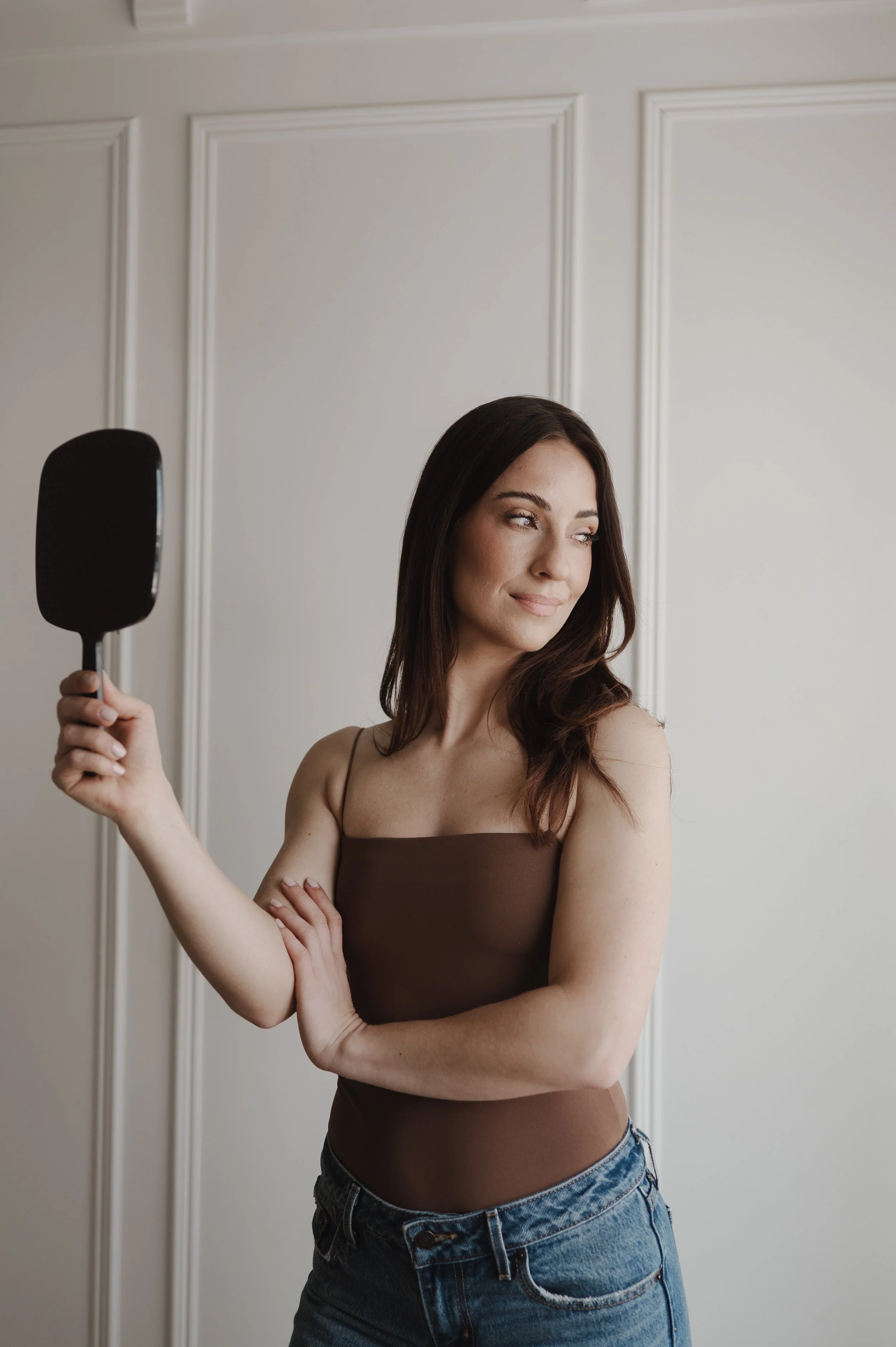 A woman with shoulder-length brown hair and light skin holding a black spatula in her right hand, standing in front of a white wall with decorative molding. She is wearing a brown tank top and blue denim jeans, and is looking to her left with a slight smile.